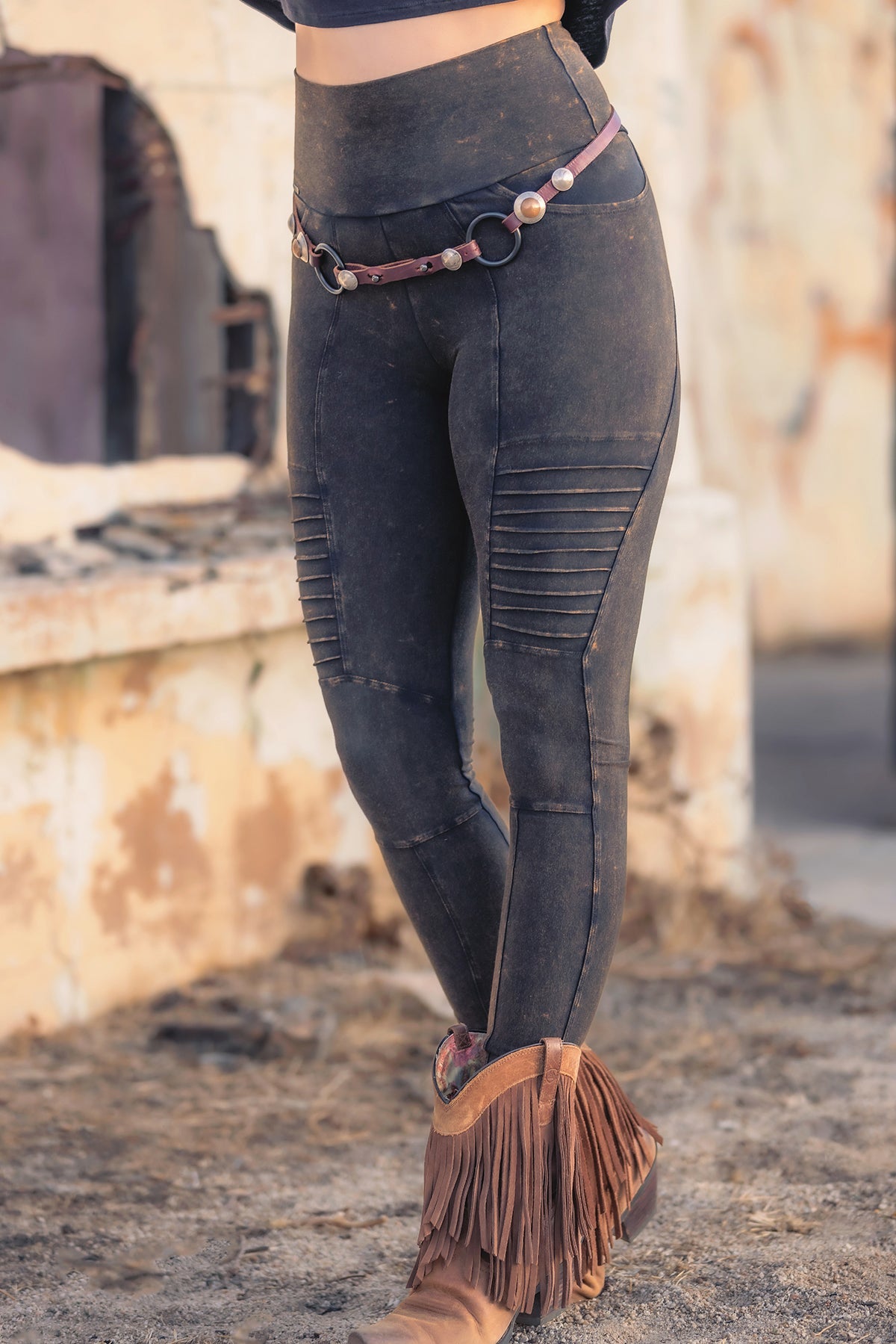 A person wearing high-waisted, dark, moto-style leggings with ribbed detailing, a decorative belt, and tan fringe ankle boots stands on a sandy ground near a weathered, light-colored wall.