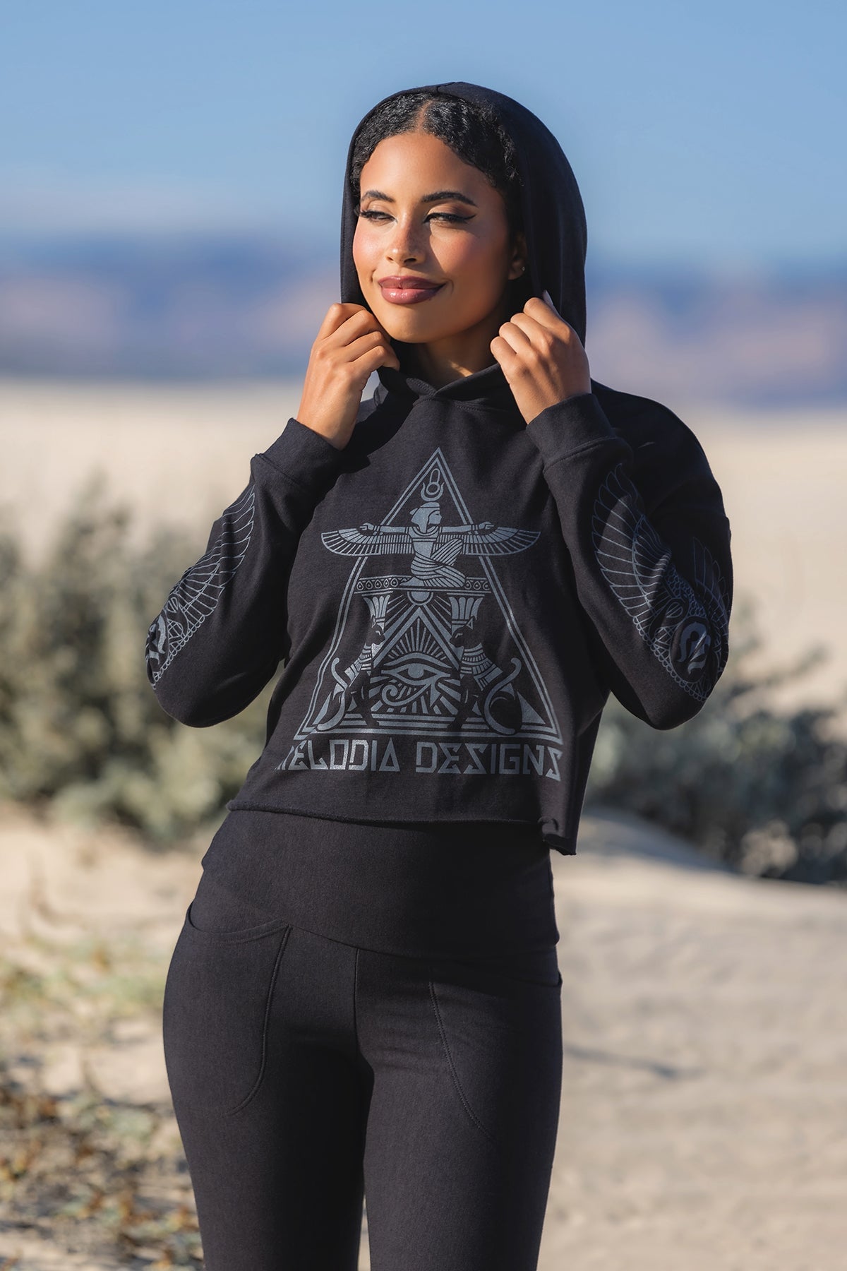 A woman stands outdoors on sand, wearing a black hoodie with an intricate geometric and bird design on the front and sleeves. She is holding the hood and smiling slightly, with plants and mountains in the background.