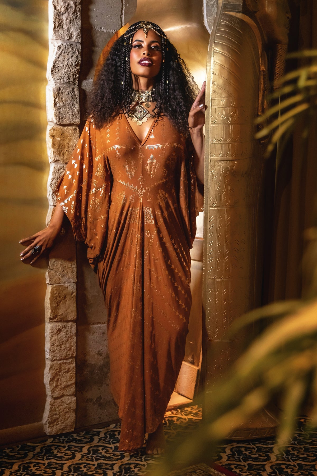 Woman with curly hair wears a purple and gold patterned dress with a high slit, large earrings, and a gold necklace, standing on a sandy background.