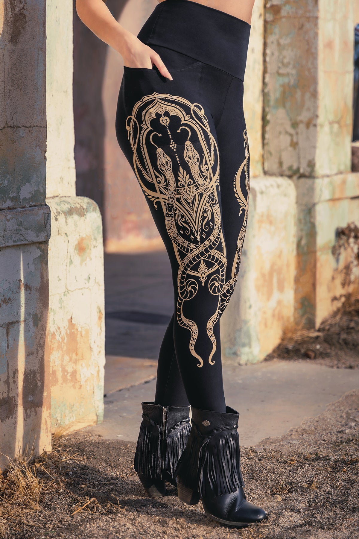 A person stands outdoors wearing black leggings with ornate gold designs and black fringed boots, next to weathered, textured stone pillars.