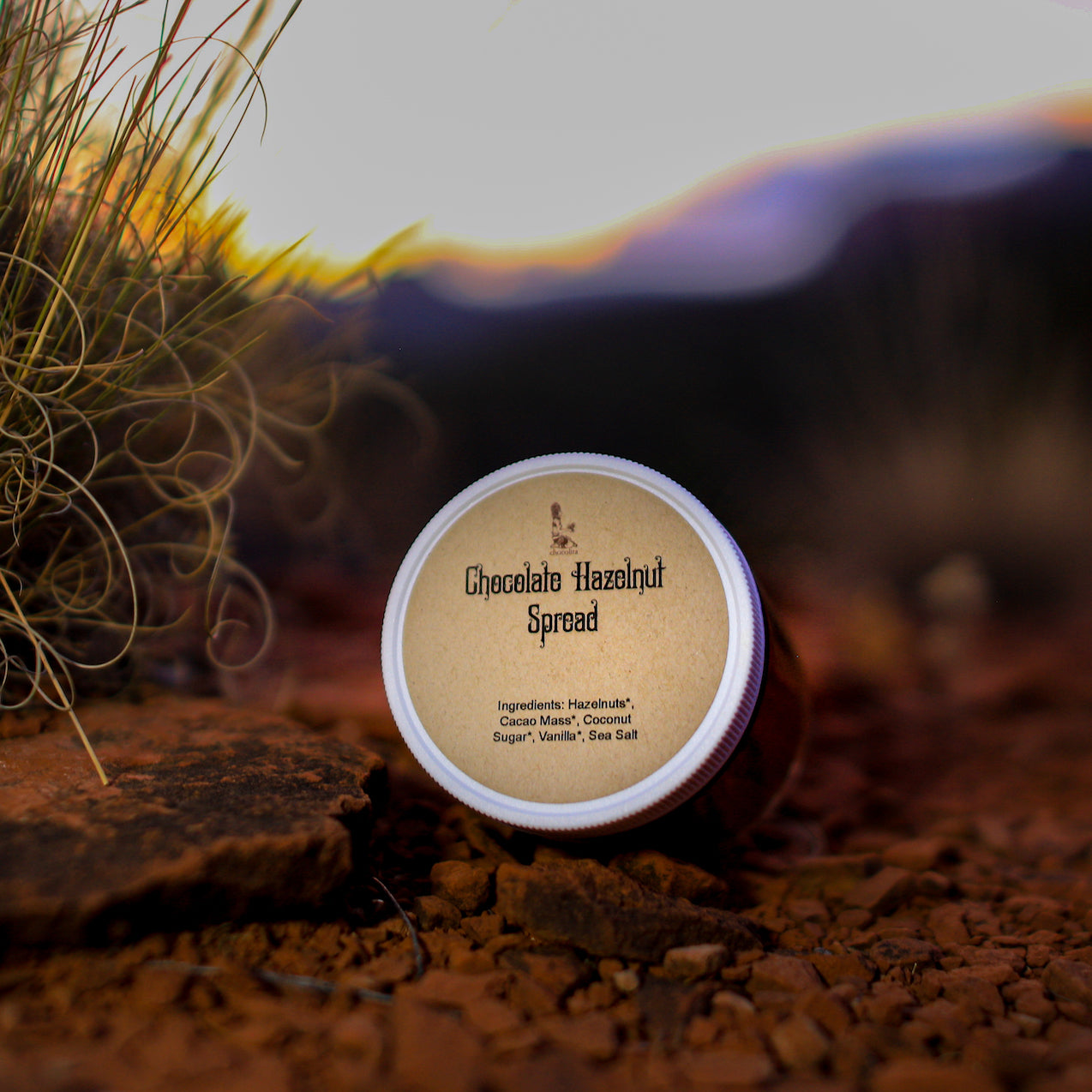 A jar labeled Hazelnut Chocolate Spread by chocolita.com sits on reddish-brown rocky ground near dry grass, with blurred mountains and a sunset behind. The label lists hazelnuts, cocoa, coconut oil, sugar, vanilla, and sea salt.