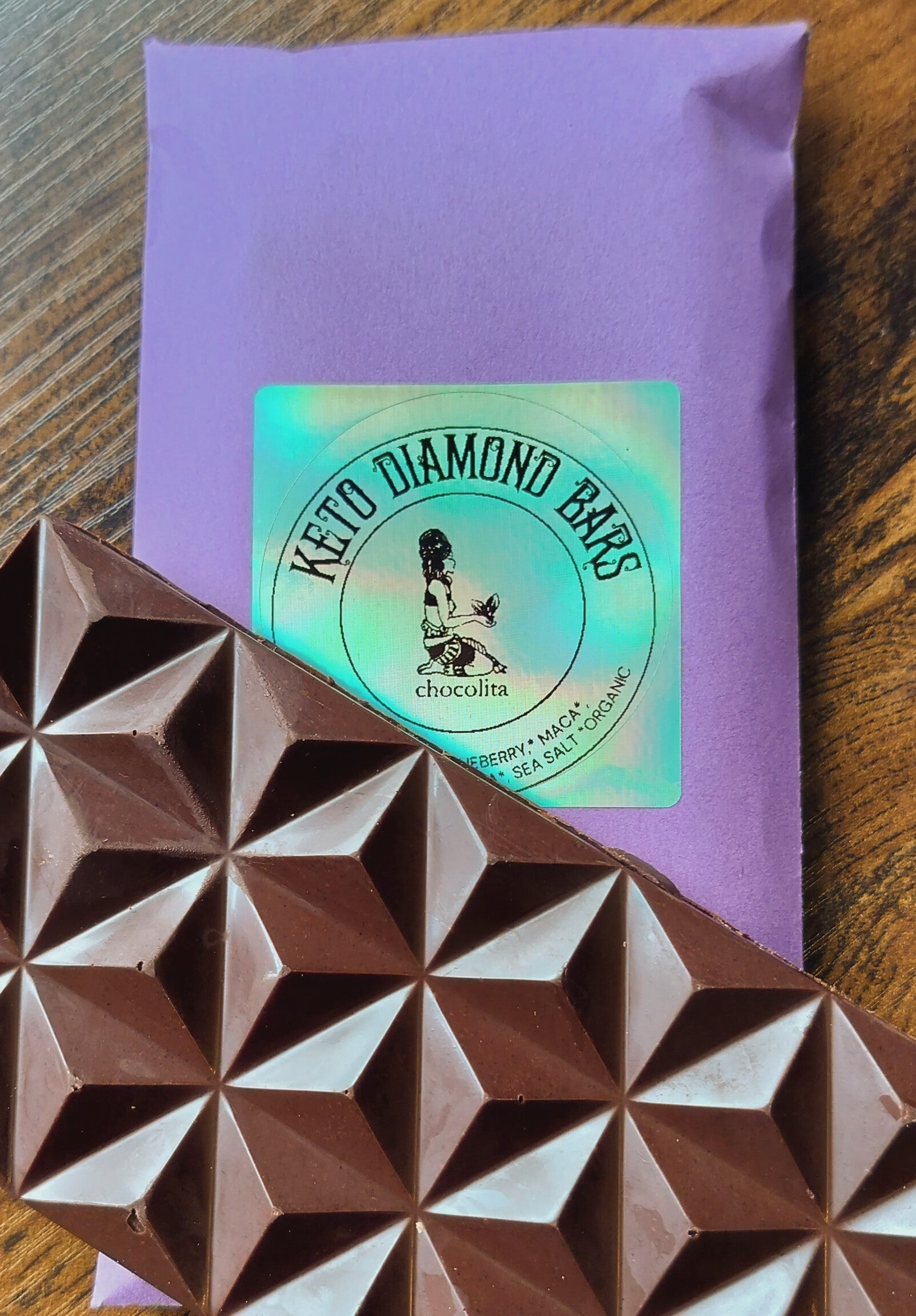A geometric raw dark chocolate bar sits on a purple chocolita.com “Keto Diamond Bar” package, featuring a holographic sticker with an illustrated figure, all arranged on a wooden surface.