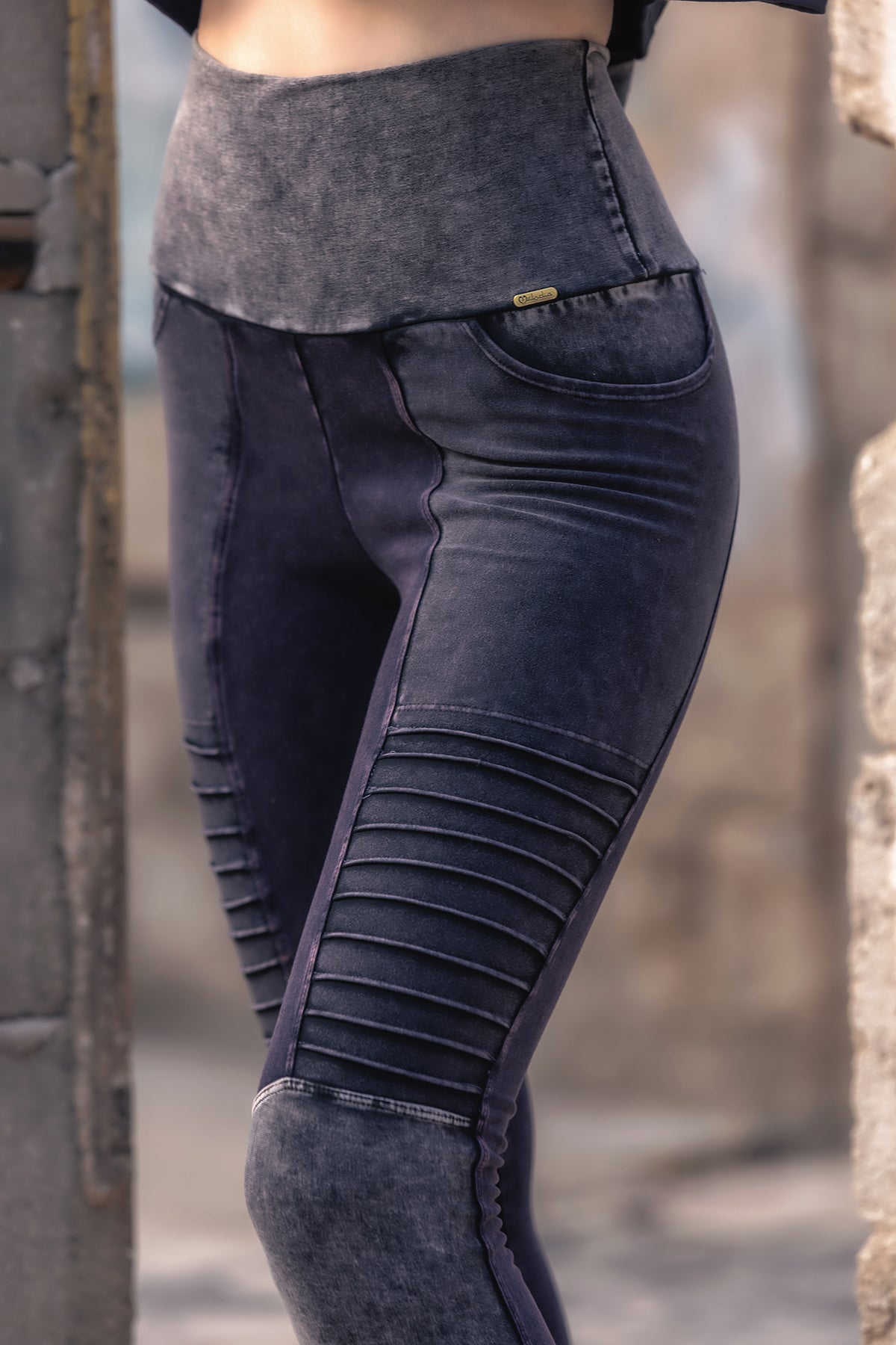 Wearing the Melodia Designs River Rock V2 Moto Xtreme Leggings—high-waisted, moto-inspired leggings crafted from dark denim with ribbed thigh detailing and visible seams—a person stands outdoors by a rough, unfinished concrete wall.