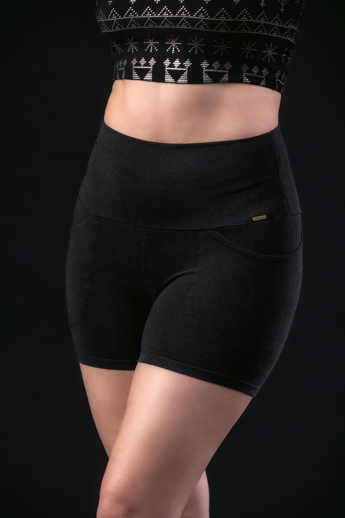 Wearing a black high-waisted crop top with a geometric pattern and Melodia Designs Alice Pocket Short, this person stands against a dark background. The face is not visible.