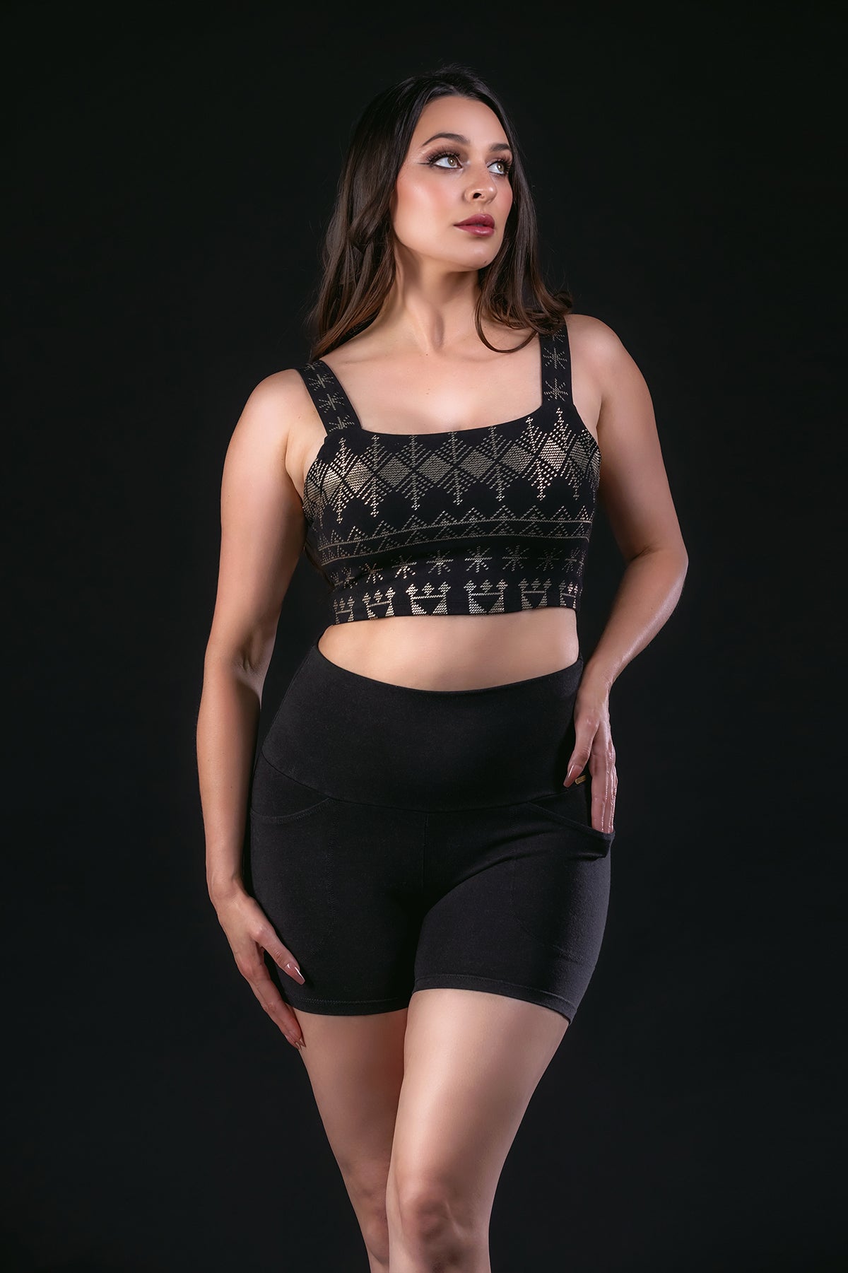 A woman poses confidently against a black backdrop, wearing Melodia Designs’ Alice Pocket Short and a black patterned crop top, with one hand on her hip and the other by her side.
