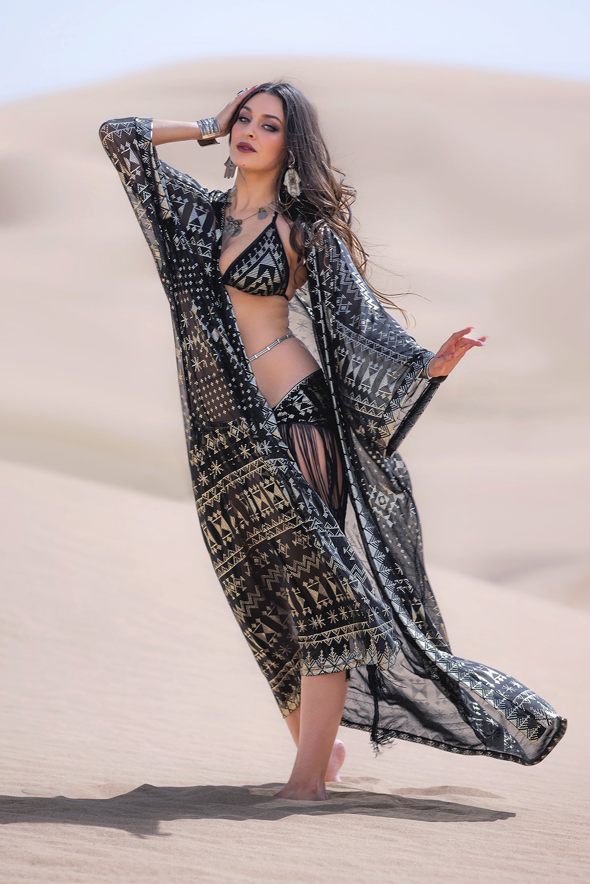 A woman stands barefoot on sand dunes, draped in the Melodia Designs Alkimiya Zsa Zsa Robe Obsidian over a fringed bikini. With long wavy hair, bold makeup, bracelets, and earrings, she channels old Hollywood glamour in this striking statement piece.