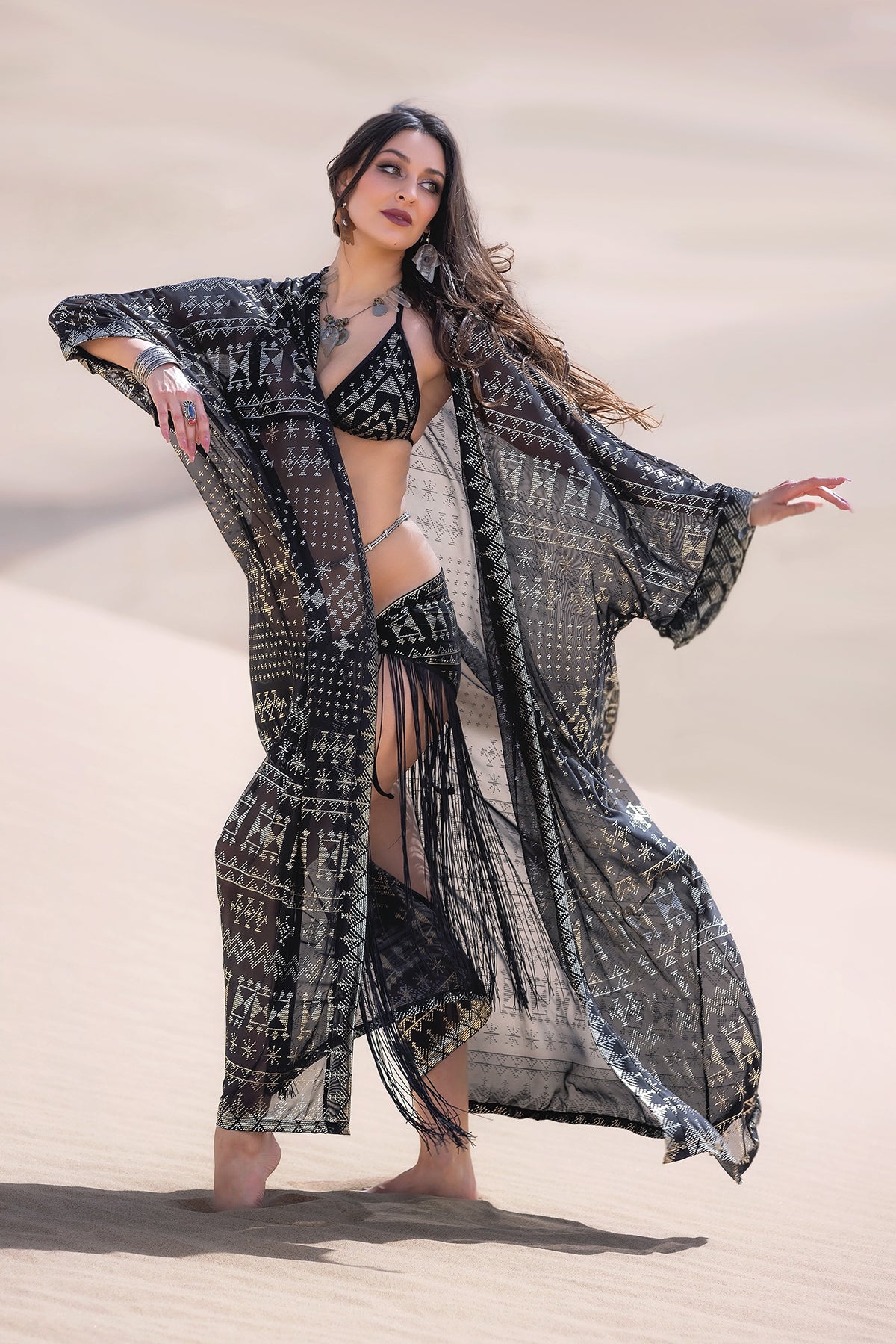 A woman poses in the desert wearing the Alkimiya Zsa Zsa Robe Obsidian by Melodia Designs over a black bikini and fringe skirt, evoking old Hollywood glamour with one arm extended amidst sand dunes.