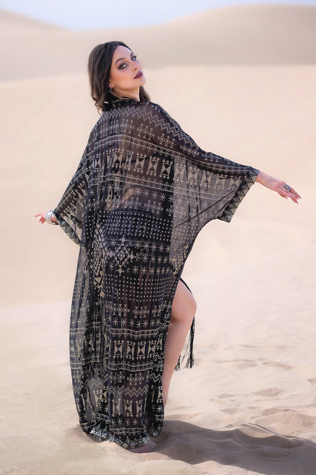 A woman poses gracefully in a sandy desert, channeling old Hollywood glamour in the Melodia Designs Alkimiya Zsa Zsa Robe Obsidian over a black outfit, looking over her shoulder with one arm extended and leg slightly raised.