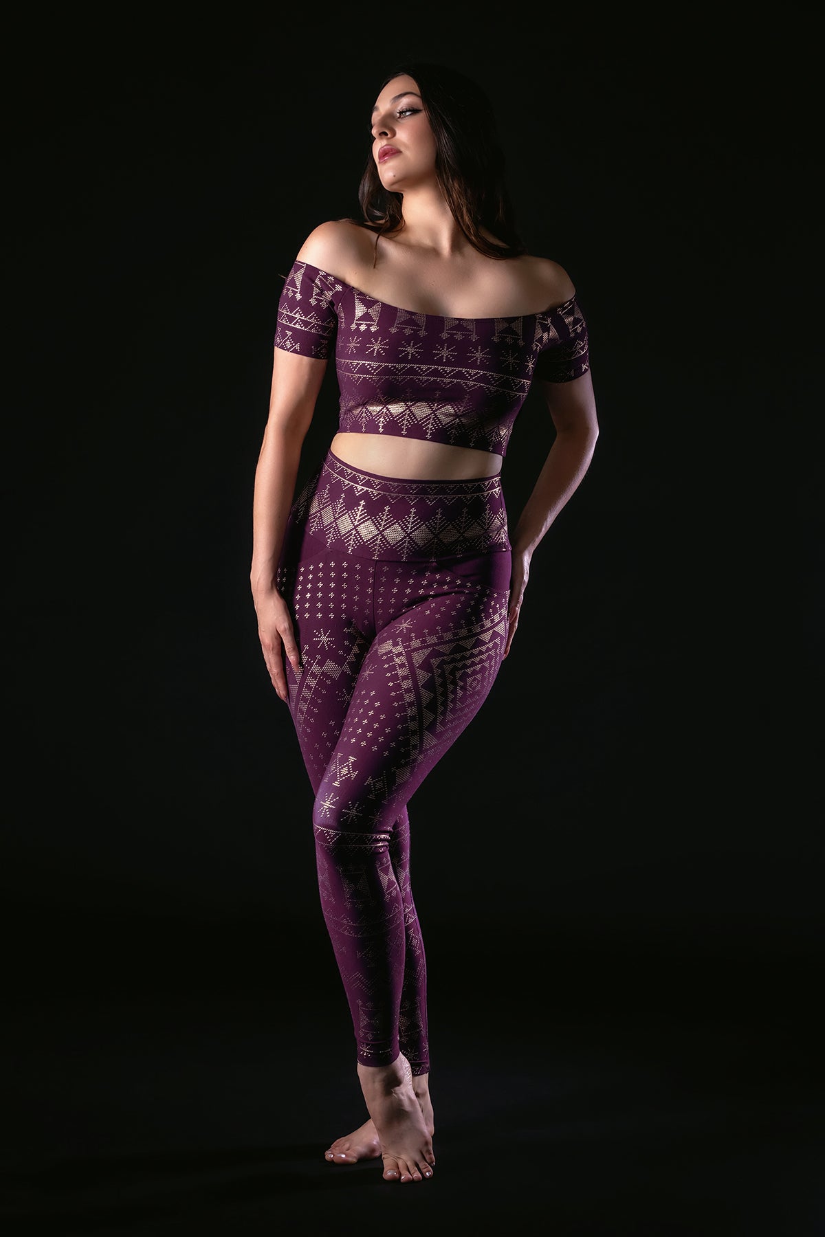 A woman poses barefoot against a black background, wearing Melodia Designs Alkimiya Elsa Legging Aubergine-Gold, a high-rise full-length legging with geometric patterns, paired with a matching purple off-the-shoulder crop top.