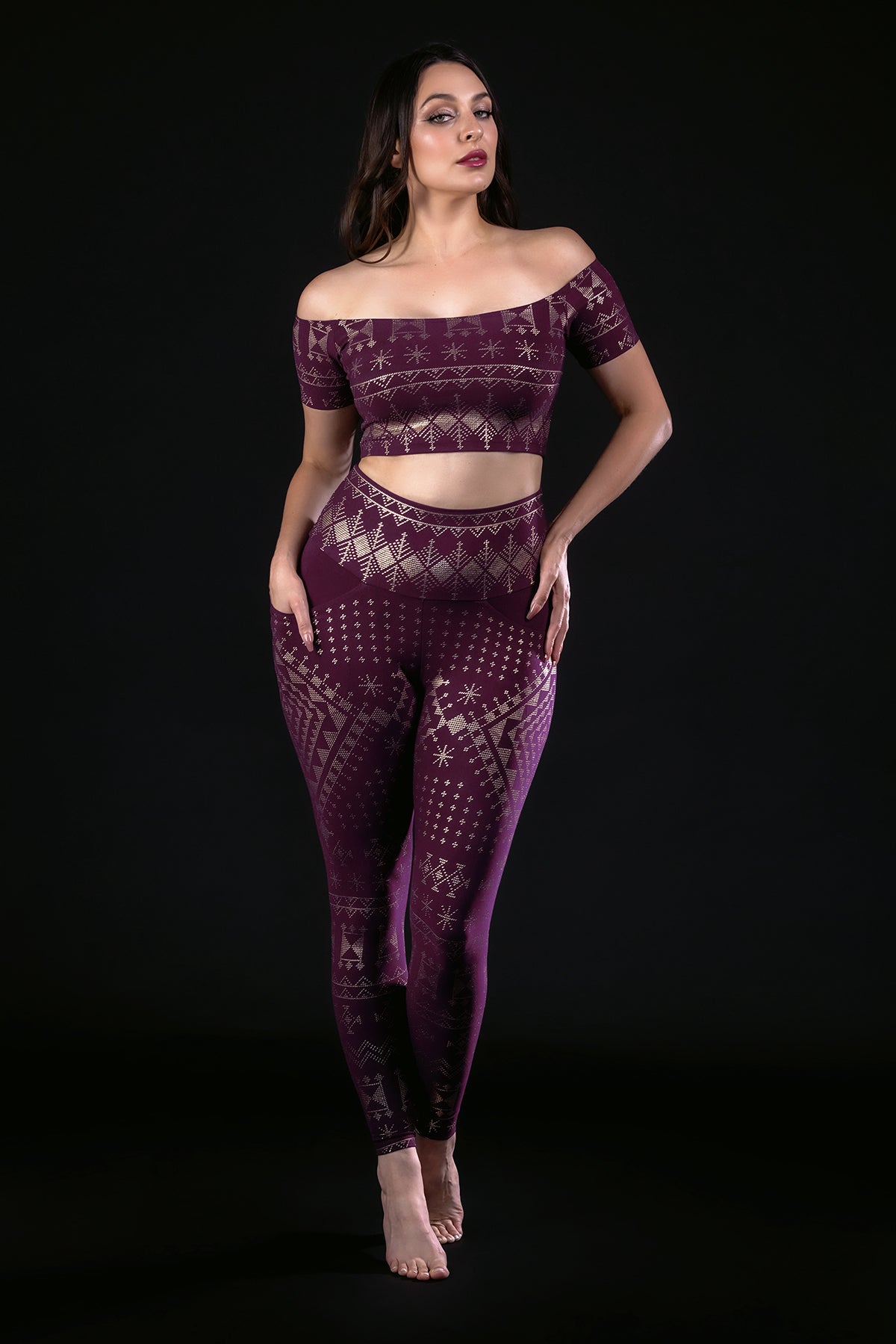 A woman poses barefoot against a black background, wearing the Melodia Designs Alkimiya Allie Crop Top Aubergine-Gold and matching leggings with gold geometric and snowflake patterns—crafted from eco-luxe fabric for stylish layering.