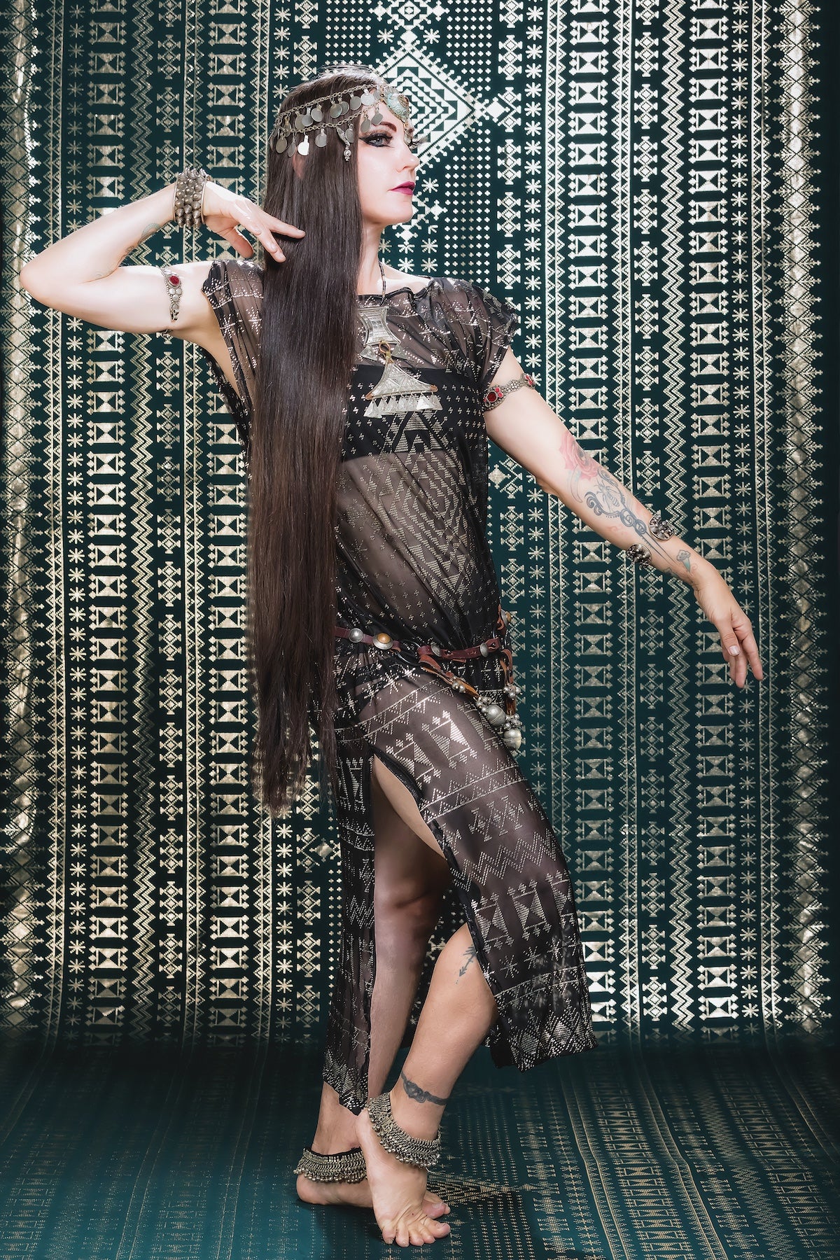 Wearing the Melodia Designs Alkimiya Nadia Dress in Obsidian-Antique Silver and a metallic headpiece, she poses gracefully against a geometric backdrop, exuding tribal artistry and effortless resort glamour.