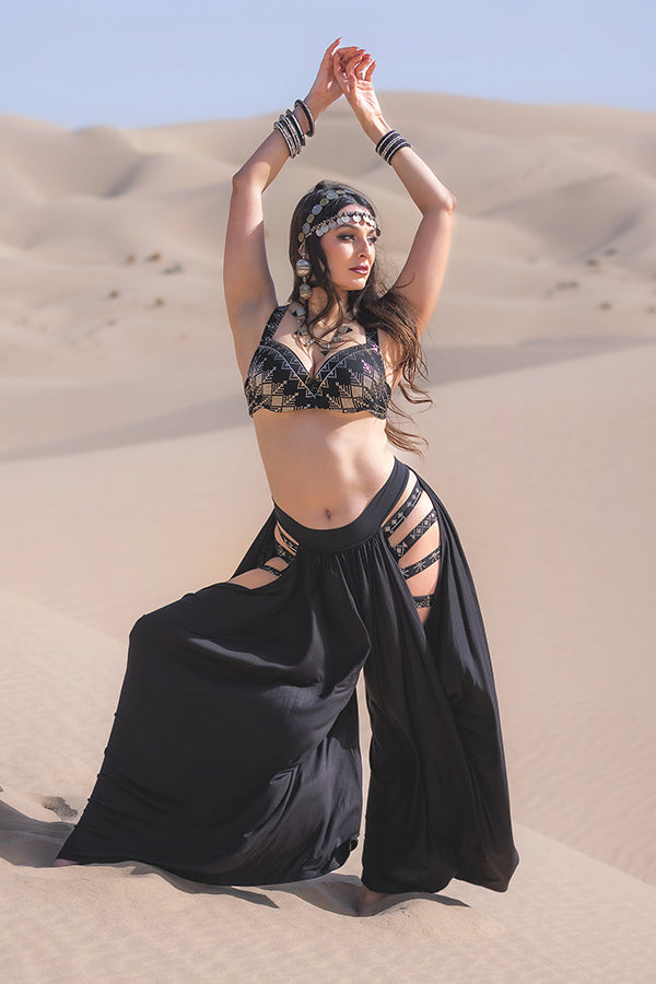 A woman poses gracefully with raised arms in a sandy desert, wearing Melodia Designs Aurora Peekaboo Palazzo Obsidian.