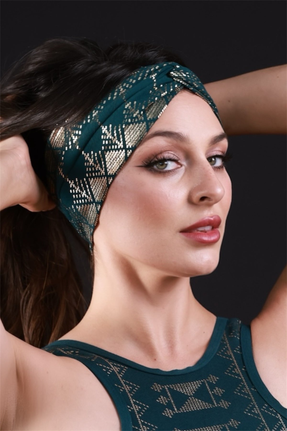 A woman with long dark hair wears the Melodia Designs Alkimiya Beauty Band Malachite—a teal and gold geometric hair accessory—paired with a matching top, posing with raised arms and turned head against a black background for an elevated look.