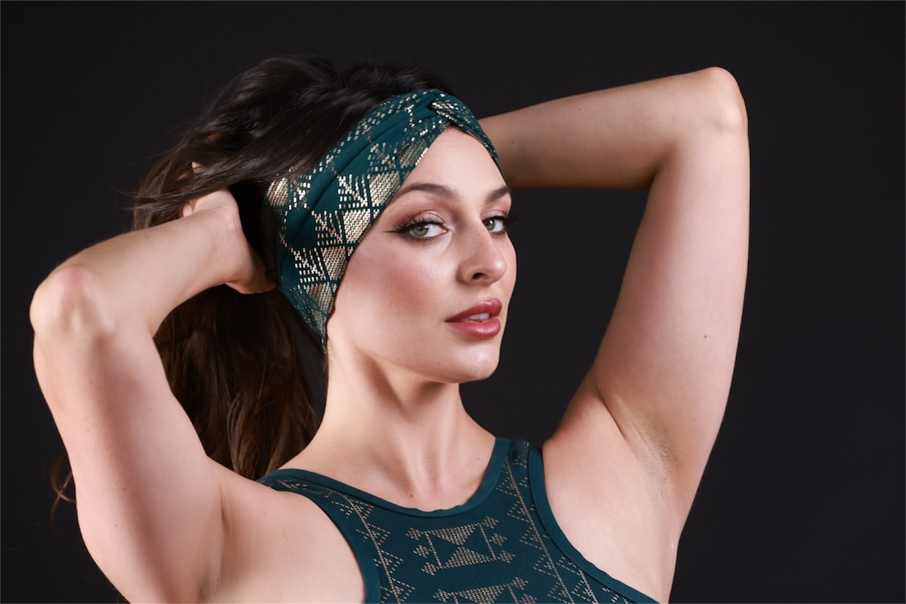 A woman with fair skin and dark hair pulls her hair back, wearing the Alkimiya Beauty Band Malachite by Melodia Designs and a matching green sleeveless top, looking confidently at the camera against a dark background.
