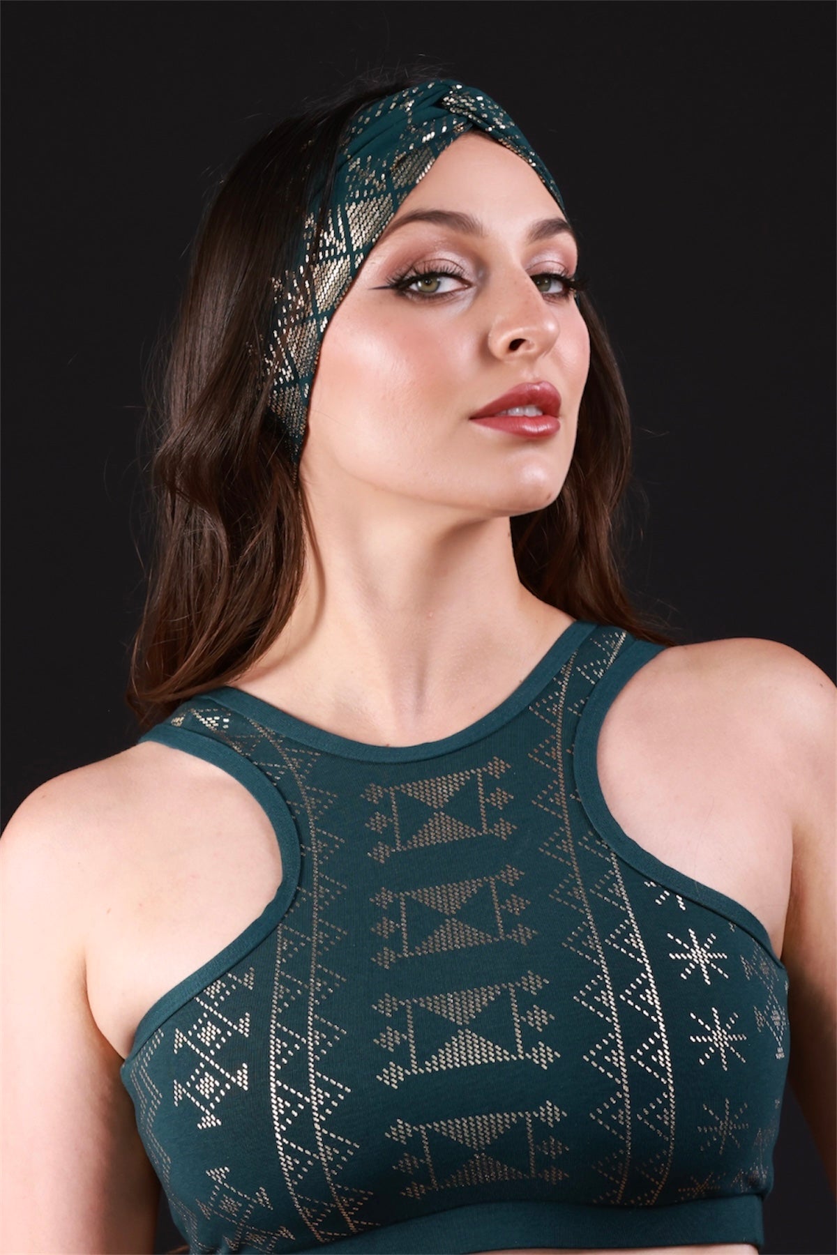 A woman with long brown hair wears the Melodia Designs Alkimiya Beauty Band Malachite and matching sleeveless top, posing confidently against a black background to showcase the brand’s elegant hair accessory design.