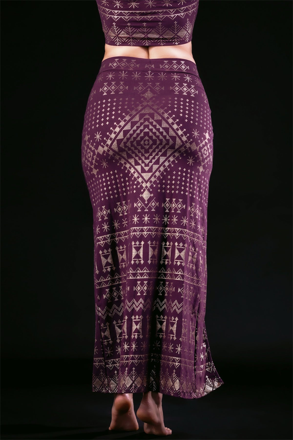 A person stands barefoot, back to the camera, wearing the Melodia Designs Alkimiya Cairo Maxi Skirt in Aubergine-Gold—a high-waisted, fitted A-line skirt featuring intricate geometric and tribal patterns on a black background.