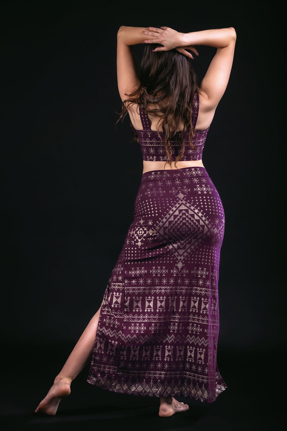 A woman stands barefoot against a black background, facing away with arms raised and crossed above her head. She wears the Alkimiya Cairo Maxi Skirt Aubergine-Gold by Melodia Designs, paired with a matching purple crop top made from eco-friendly fabric.