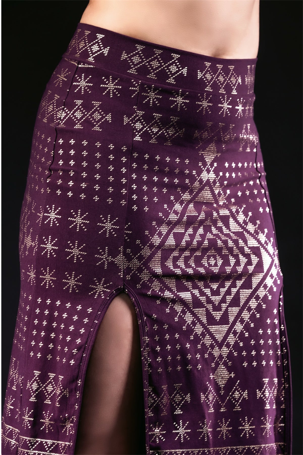 A close-up of a person wearing the Alkimiya Cairo Maxi Skirt in Aubergine-Gold by Melodia Designs, featuring an A-line silhouette, high waist, gold geometric patterns, thigh-high slit, and eco-friendly fabric against a black background.