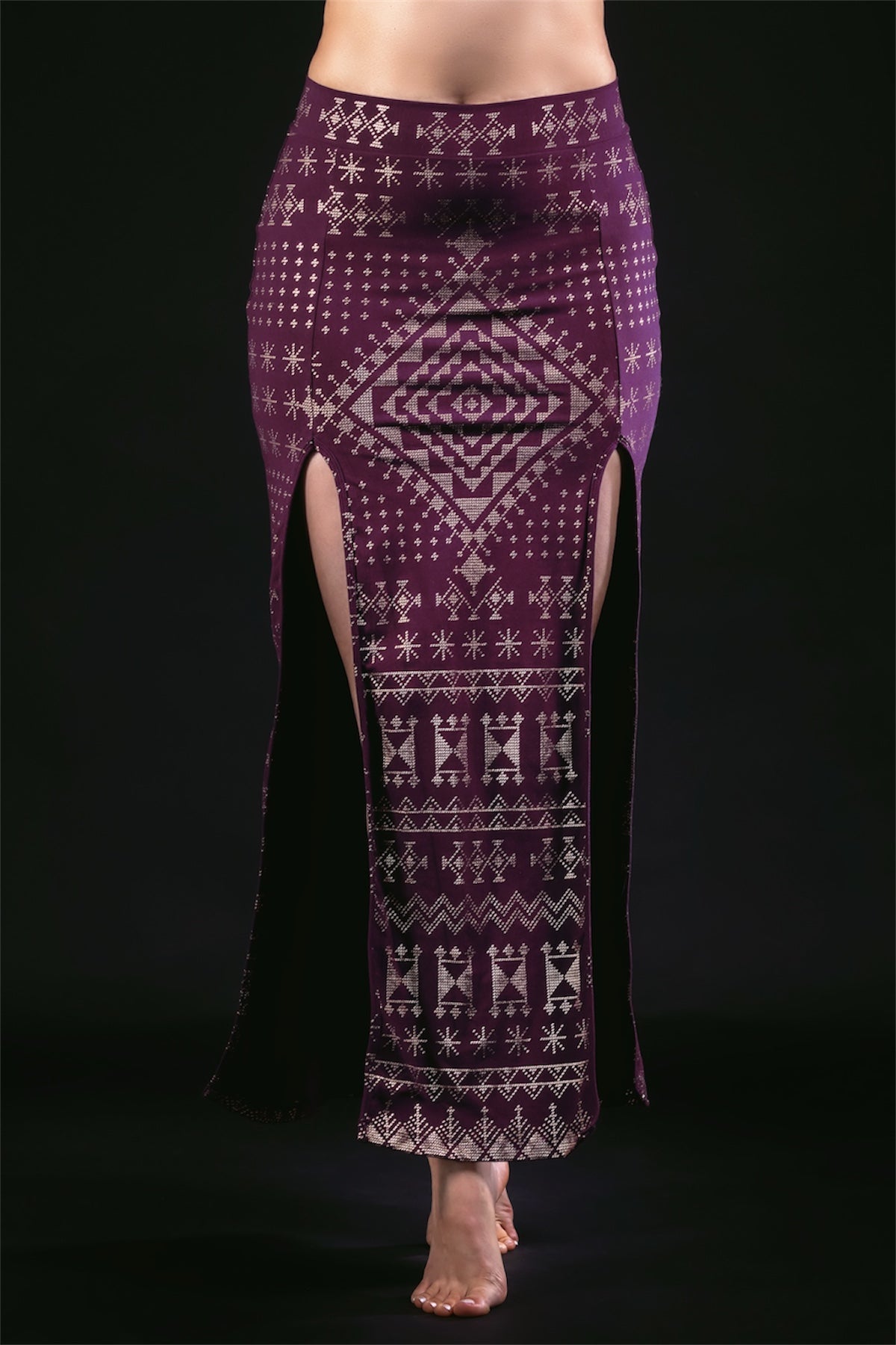 A person stands barefoot against a black background wearing the Melodia Designs Alkimiya Cairo Maxi Skirt in Aubergine-Gold, a high-waisted A-line eco-friendly skirt with geometric and tribal patterns and two high front slits.