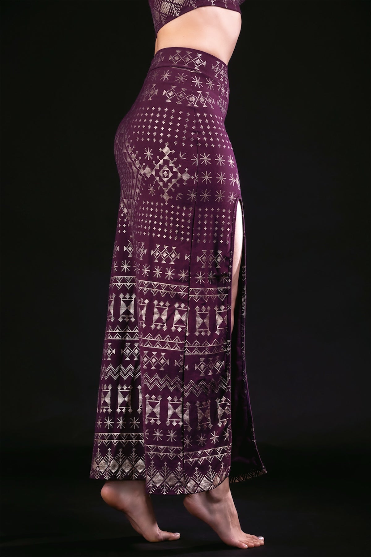 A model showcases the Melodia Designs Alkimiya Cairo Maxi Skirt in Aubergine-Gold—an A-line, high-waisted, patterned purple skirt with a side slit—posed on tiptoe against a black background, showing only the lower torso and legs.