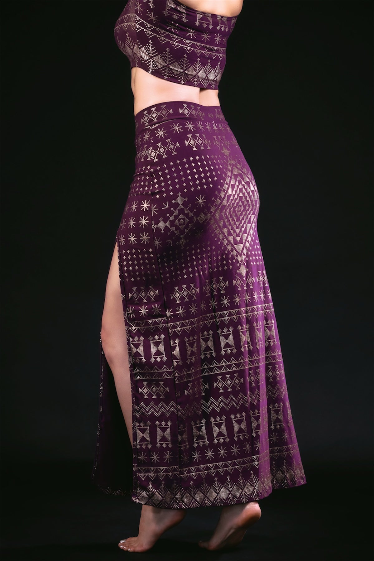 A person poses barefoot against a black background, wearing the Melodia Designs Alkimiya Cairo Maxi Skirt in Aubergine-Gold with a high slit and gold geometric patterns. Their face is not visible.