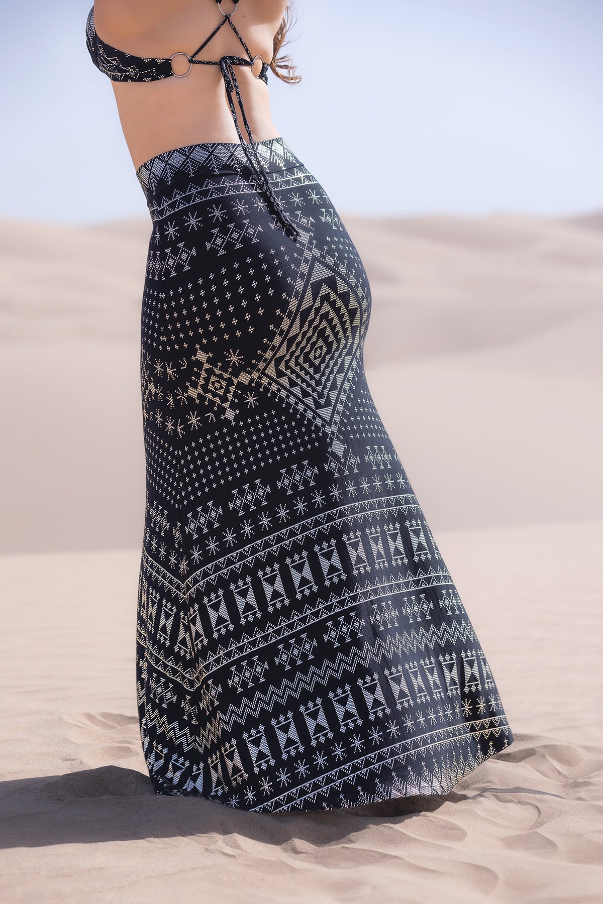 A person stands in a sandy desert under a clear sky, wearing a black bikini top and the Melodia Designs Alkimiya Cleopatra Maxi Skirt Obsidian featuring white geometric patterns and an A-line silhouette.