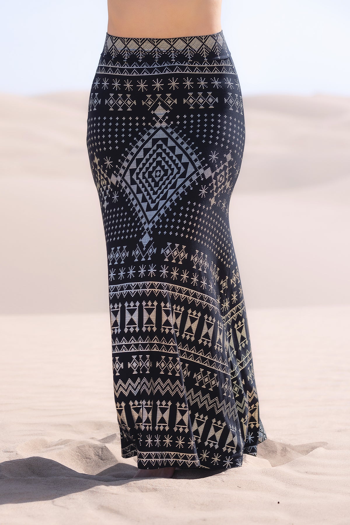 A person stands on desert sand wearing the Melodia Designs Alkimiya Cleopatra Maxi Skirt Obsidian, featuring white geometric and tribal patterns on black fabric. The flattering A-line silhouette is visible, with only the lower body shown.