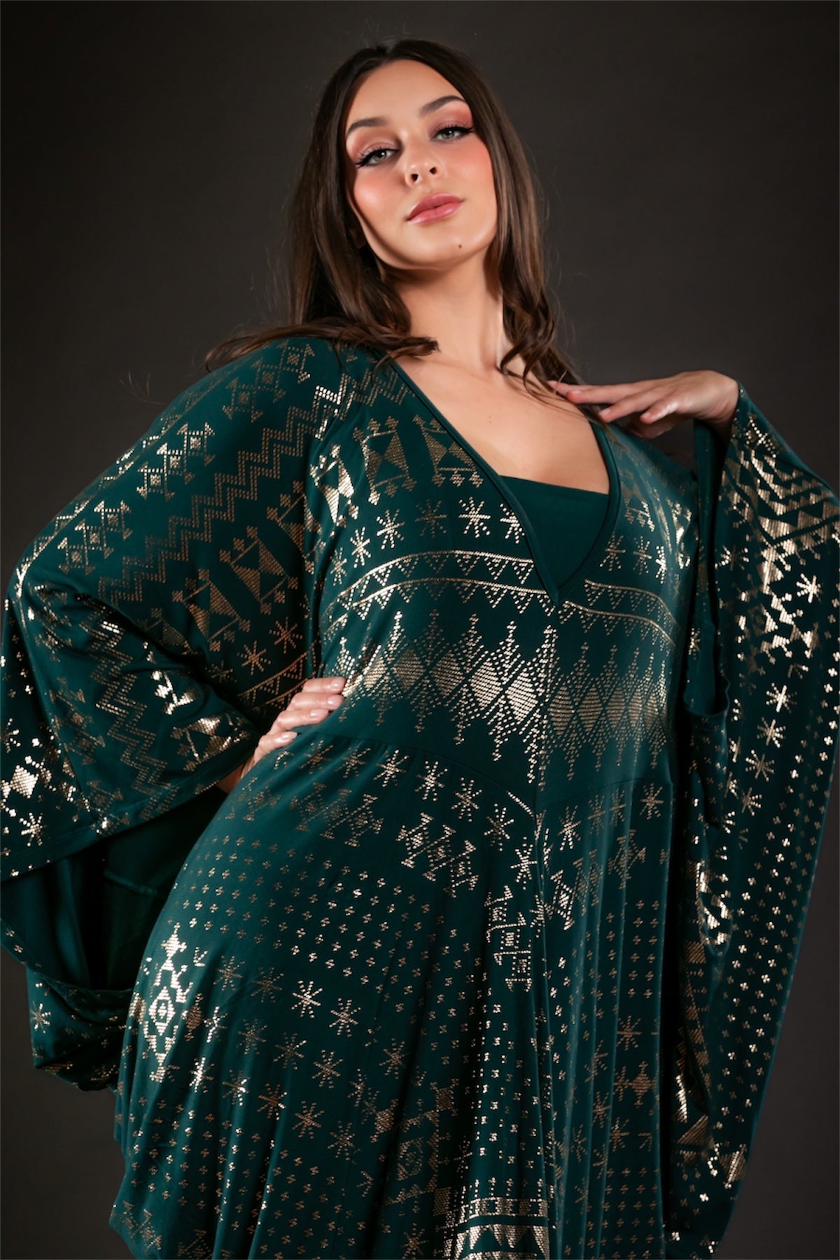 A woman poses confidently against a dark background wearing the Melodia Designs Alkimiya Contessa Caftan Malachite, a dark green dress with batwing sleeves and gold geometric patterns.