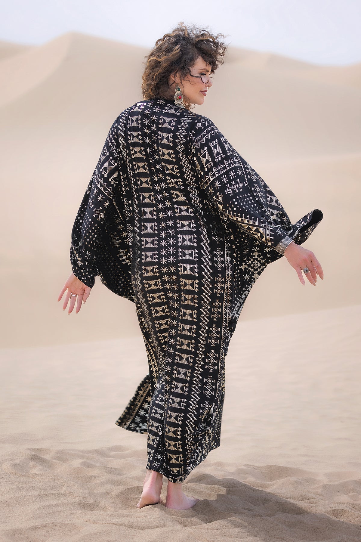 A woman with curly hair walks barefoot on sand dunes, wearing the Alkimiya Contessa Caftan Obsidian by Melodia Designs. She looks over her shoulder as the eco-luxe fabric billows around her in the gentle breeze.