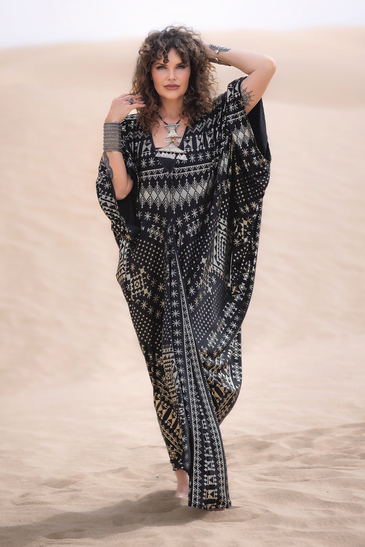 A woman with curly hair stands barefoot on sand in the Alkimiya Contessa Caftan Obsidian by Melodia Designs. The black and gold batwing-sleeved caftan and silver jewelry enhance her elegant look against the desert landscape.