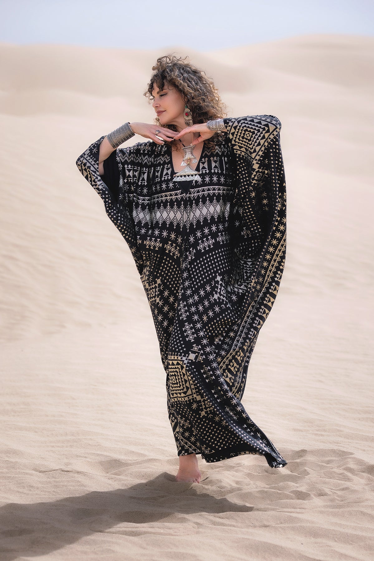 A woman stands barefoot in sand dunes, wearing the Alkimiya Contessa Caftan Obsidian by Melodia Designs. With arms raised gracefully and eyes closed, her curly hair frames her face against a sunny desert backdrop.