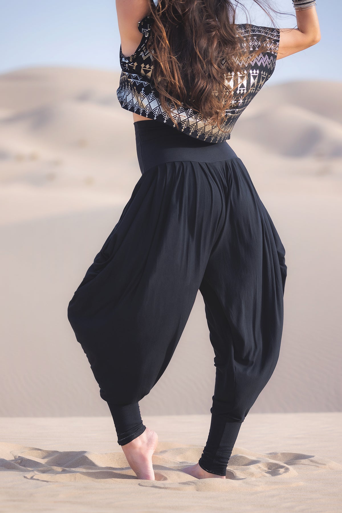 Amid sweeping sand dunes, a barefoot figure wears Melodia Designs Jazmine Harem Pant in Obsidian—eco-luxe, flowy, pleated black pants—paired with a patterned sleeveless top and long hair cascading down their back.