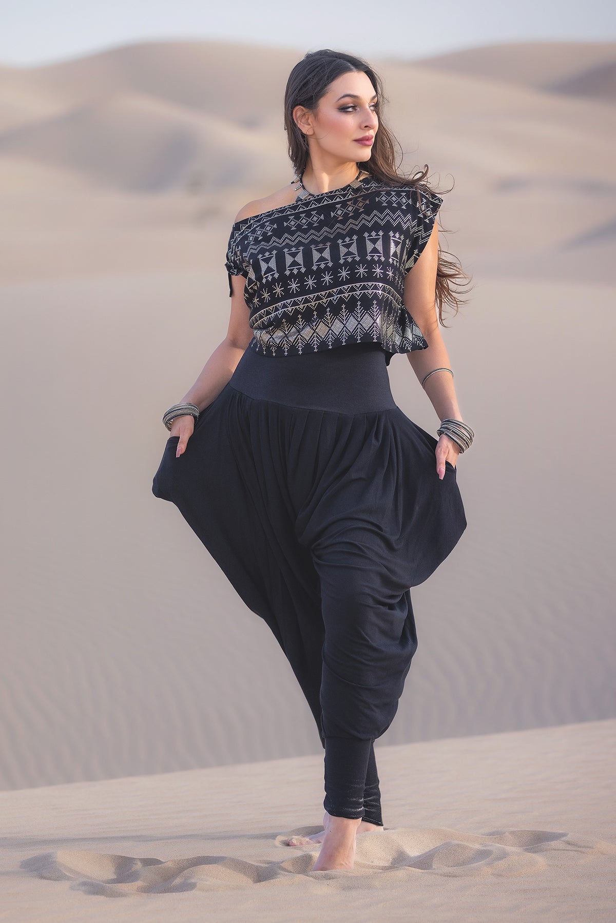 A woman stands barefoot on sand dunes, wearing Melodia Designs Jazmine Harem Pant in Obsidian and a patterned black one-shoulder top. With long brown hair, she gazes to the side, hands in her pockets.