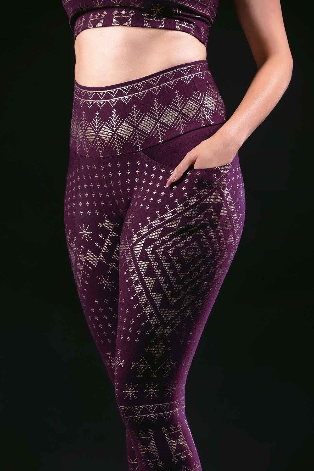 Wearing Melodia Designs Alkimiya Elsa Legging in Aubergine-Gold, featuring geometric and snowflake patterns on eco-friendly fabric, a person poses confidently with one hand on their hip against a black background.