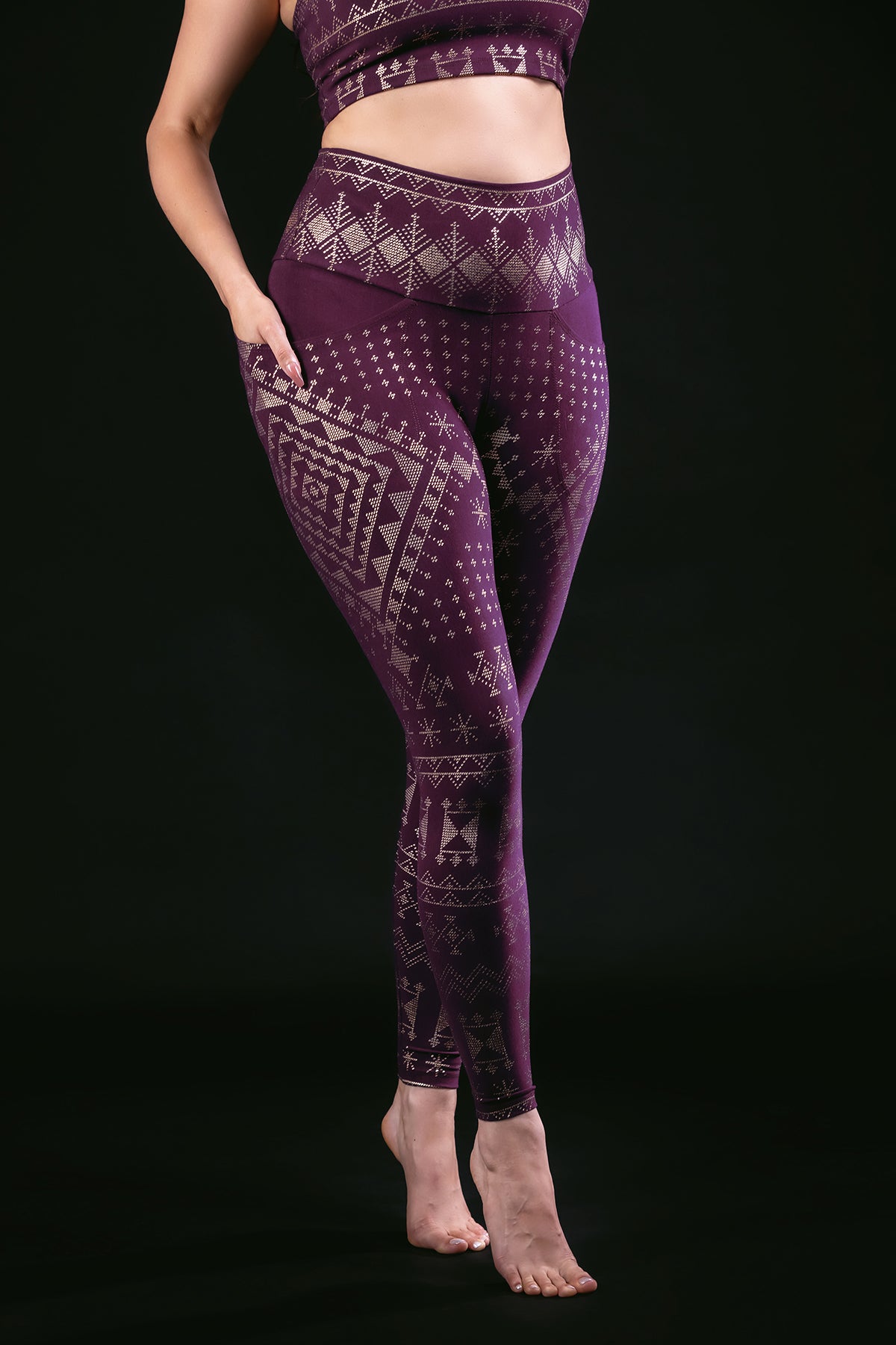 A woman stands barefoot against a black background, wearing Melodia Designs Alkimiya Elsa Legging Aubergine-Gold with a high-rise waistband and eco-friendly fabric. Her hands are on her hips, and her head is cropped out of the frame.