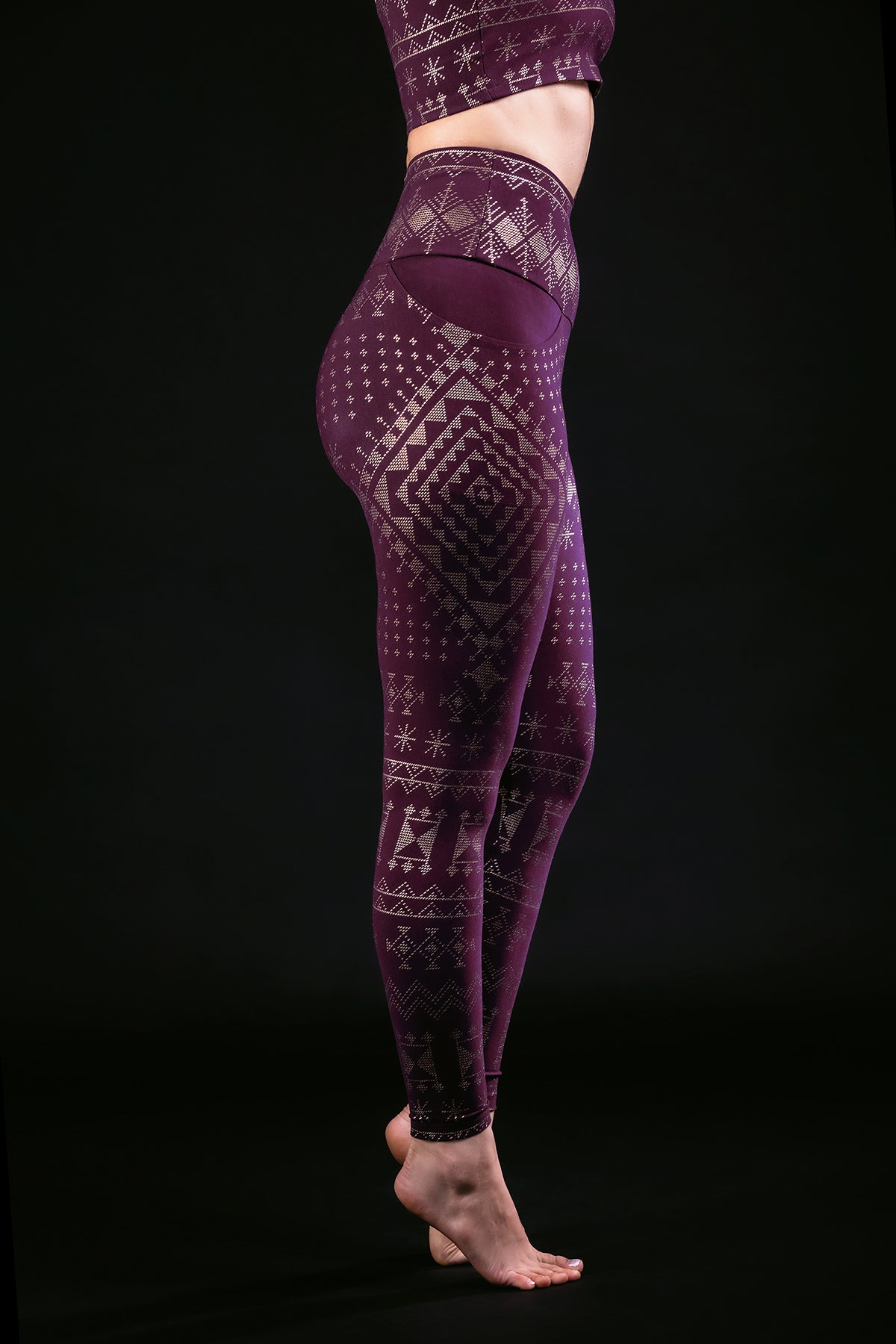 A person stands barefoot against a black background, modeling Melodia Designs Alkimiya Elsa Legging Aubergine-Gold with geometric and snowflake patterns, paired with a matching crop top. The side view features the high-rise waistband, shown from shoulders to feet.