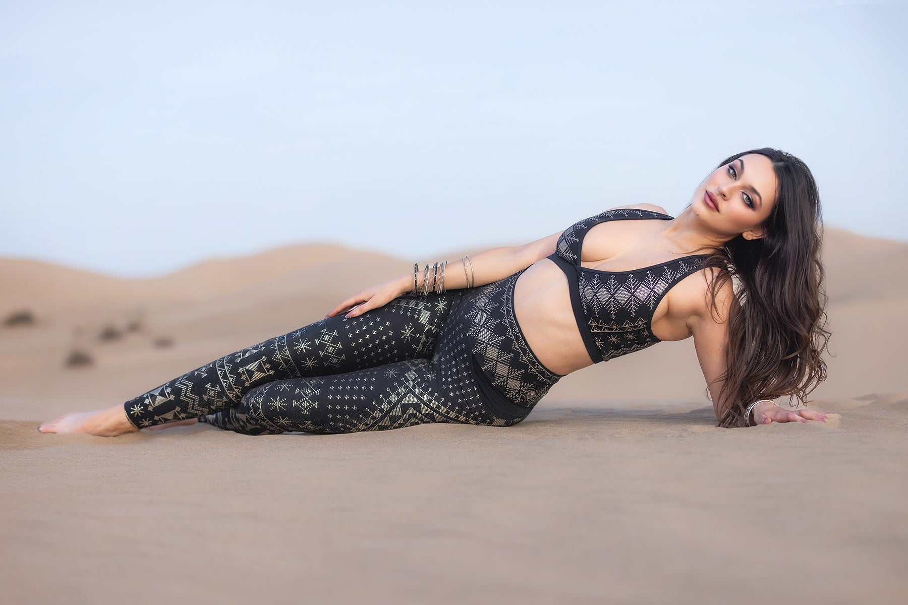 A woman with long hair, wearing Melodia Designs’ Alkimiya Elsa Pocket Legging Obsidian and a patterned sports bra, reclines on desert sand with distant dunes behind her, gazing at the camera.