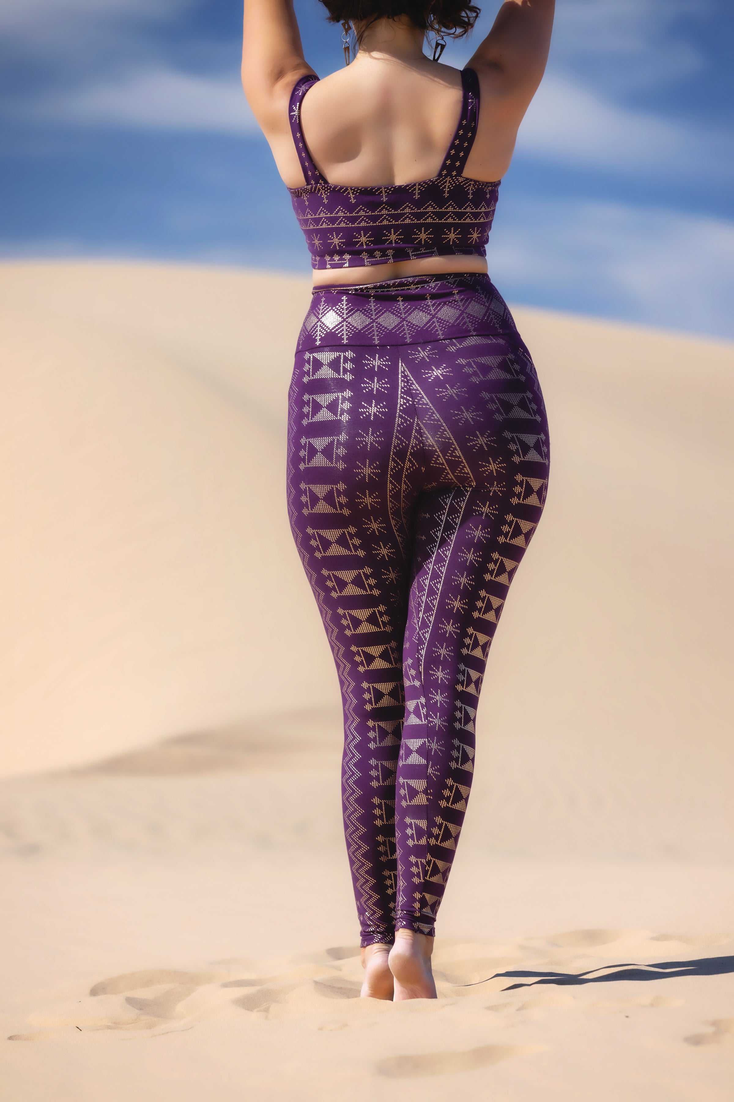 A person stands barefoot on sand dunes under a blue sky, arms raised and shown from behind, wearing Melodia Designs’ Alkimiya Elsa Legging Aubergine-Gold v2 with a matching purple sports bra.