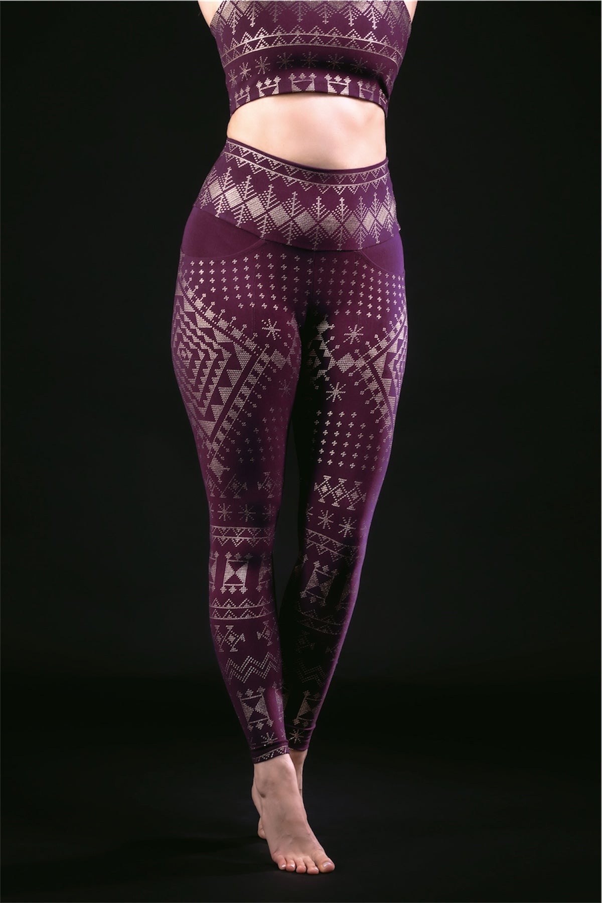 A person stands barefoot against a black background, wearing Melodia Designs Alkimiya Elsa Legging in Aubergine-Gold, featuring a high-rise waistband and white geometric and snowflake patterns. The matching crop top is partially visible; head and feet are not shown.