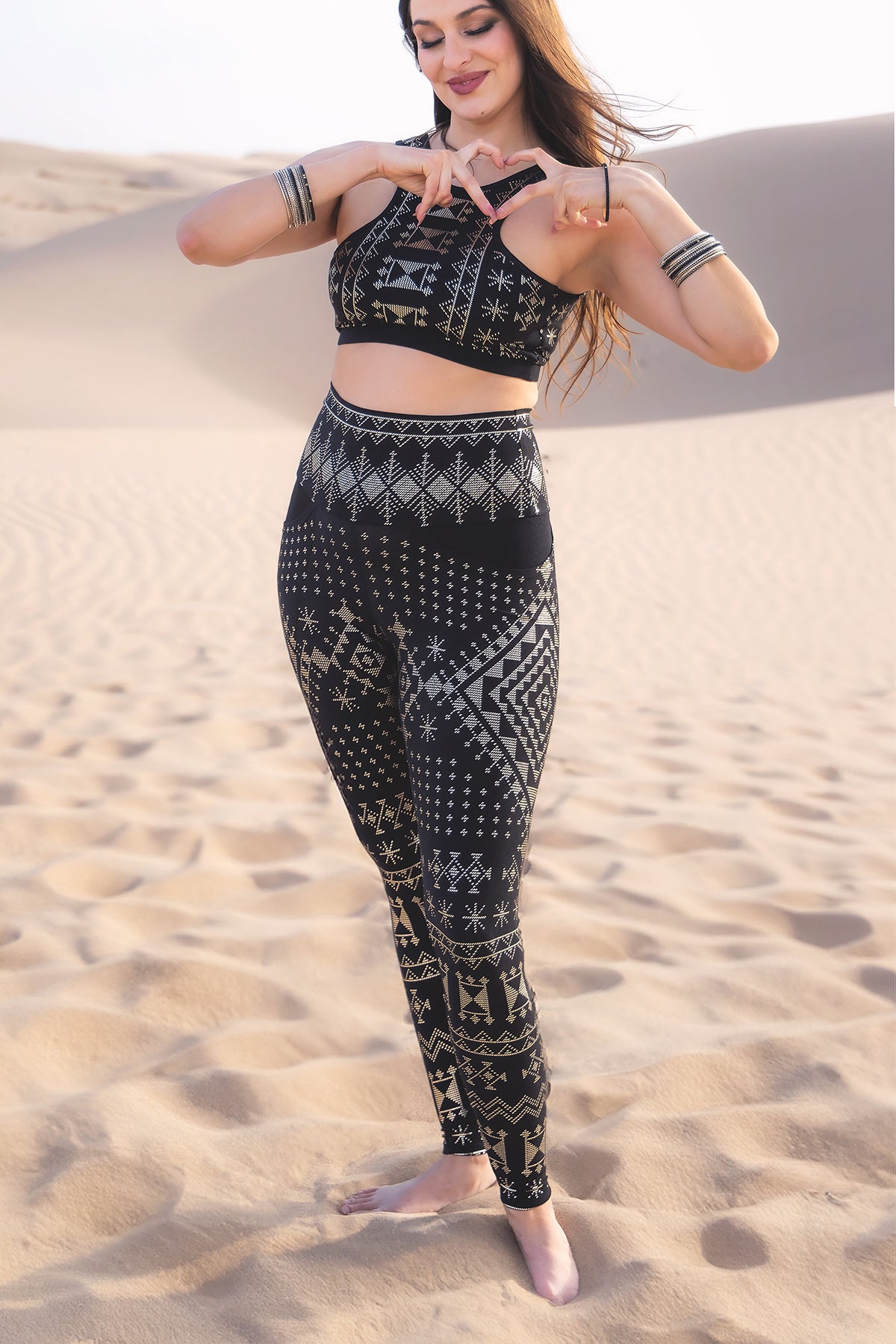 A woman stands barefoot on sand dunes, smiling as the wind blows her hair. She wears Melodia Designs Alkimiya Elsa Pocket Legging Obsidian and a matching sports bra with black and beige geometric patterns, posing confidently with her hands.
