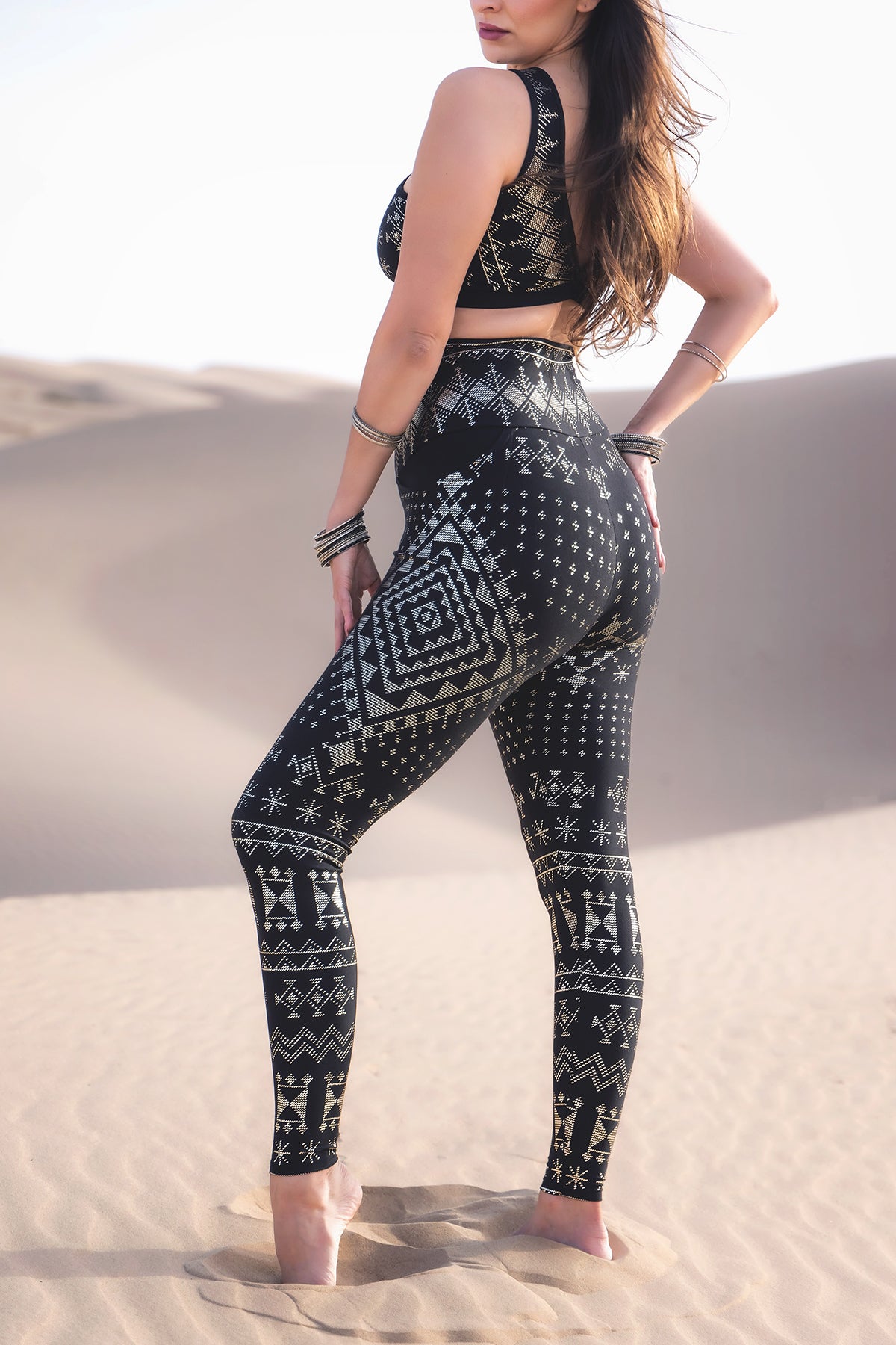 A woman with long brown hair stands barefoot on sand dunes, wearing Melodia Designs Alkimiya Elsa Pocket Legging Obsidian and a matching geometric sports bra, both crafted from eco-lux fabric, accessorized with bracelets.