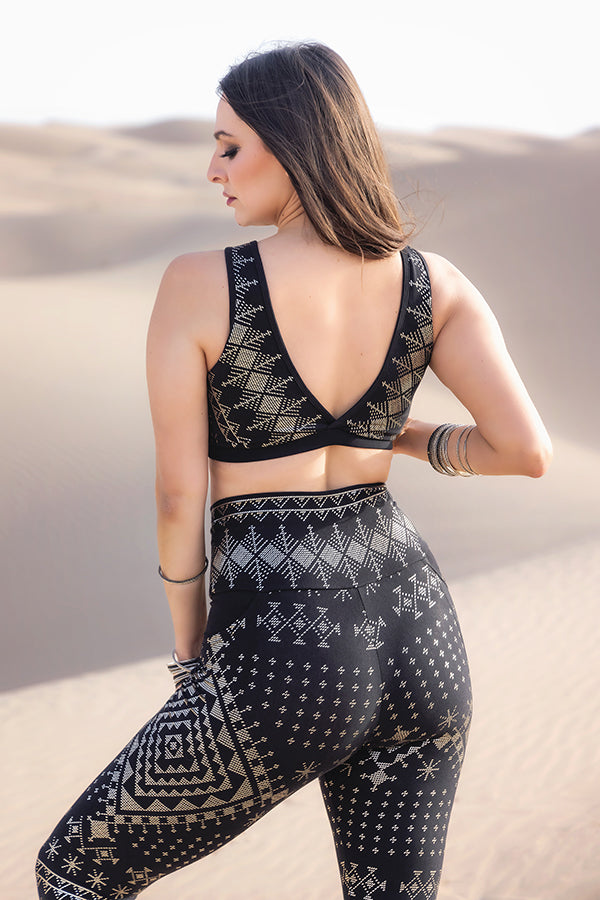 A woman with her hair down and bangles on her wrist stands in a desert, wearing the Melodia Designs Alkimiya Rainah Bra Obsidian with matching high-waisted leggings. Sand dunes extend behind her.