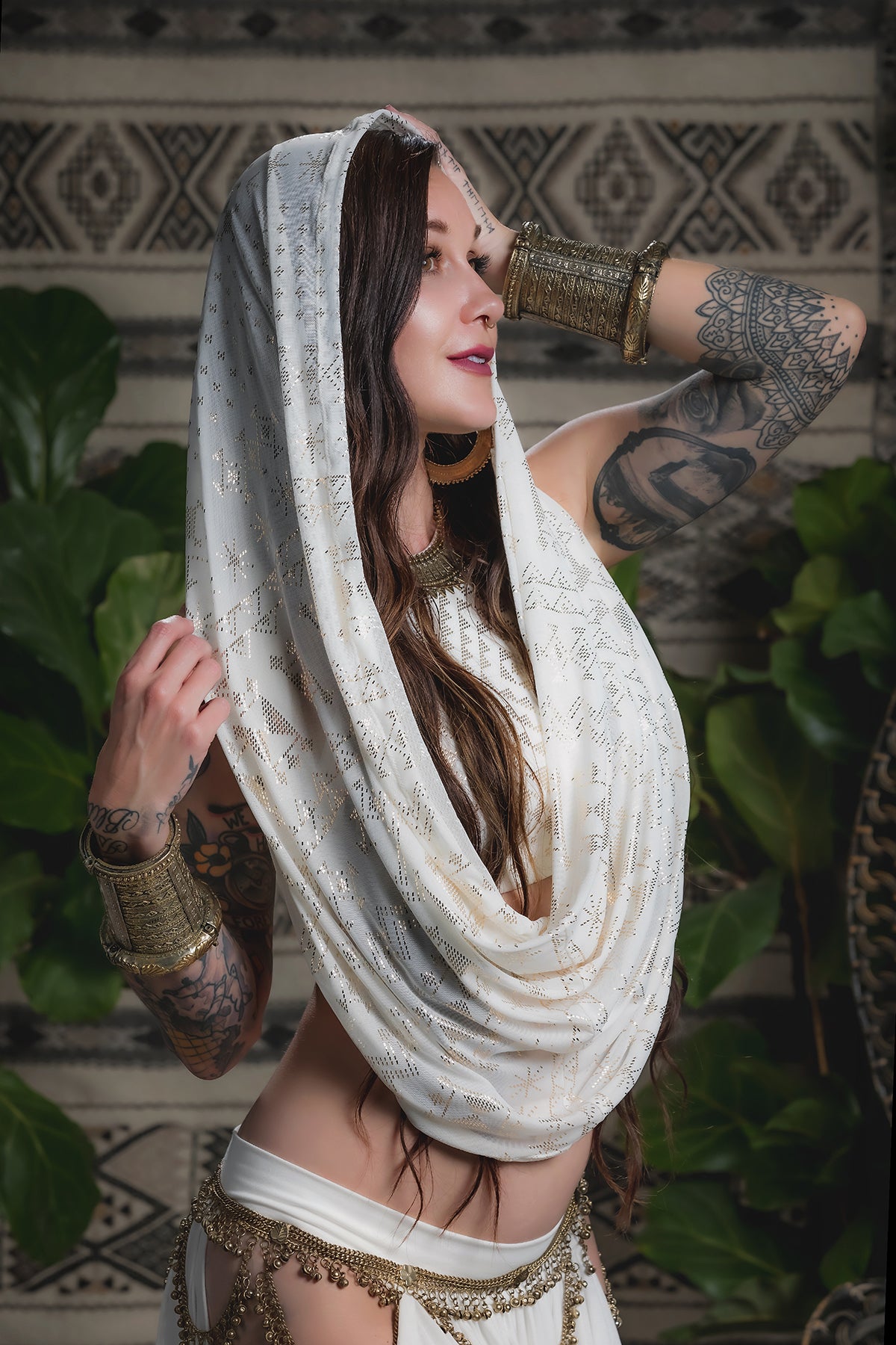 A woman models the Alkimiya Infinity Scarf Ivory by Melodia Designs, draped over her head with gold jewelry and tattooed arms, posing gracefully amidst green plants and a patterned backdrop.