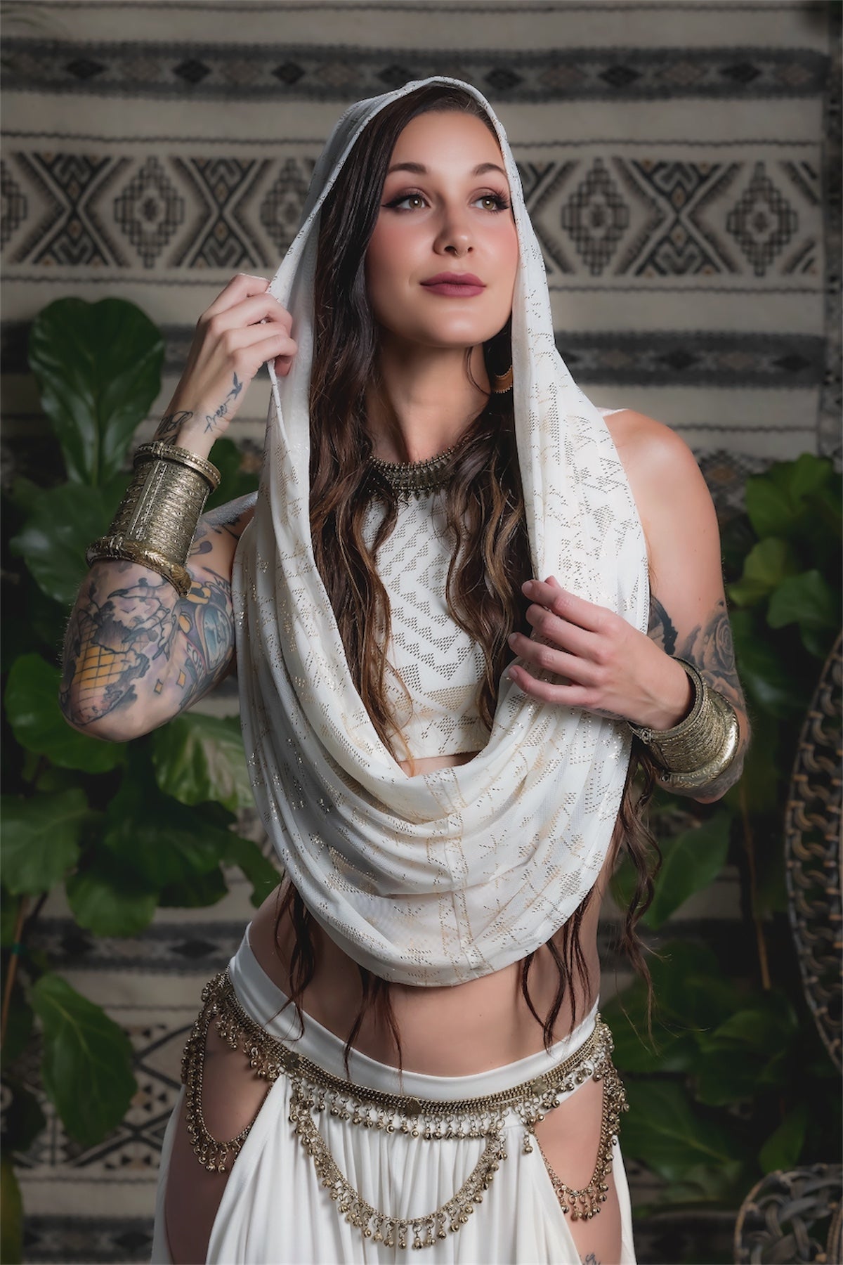 A woman with long brown hair and tattooed arms wears the Alkimiya Infinity Scarf Ivory by Melodia Designs, styled with flowing garments and chunky gold jewelry, posing before a patterned textile and green plants.