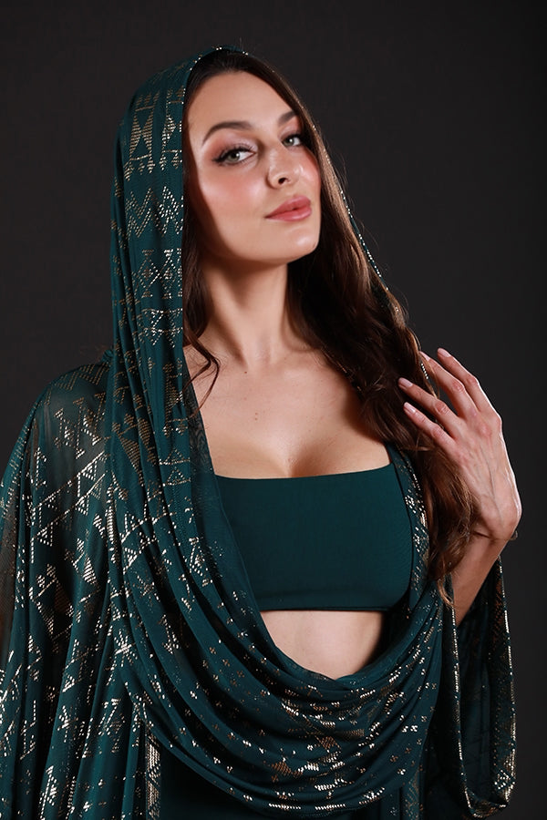 A woman confidently poses against a dark background, wearing the Melodia Designs Alkimiya Infinity Scarf Malachite in deep green and gold over her head and shoulders, paired with matching green attire. Her hand is raised near her face as she gazes at the camera.