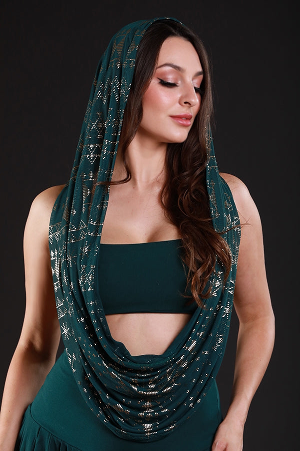 A woman models the Alkimiya Infinity Scarf Malachite by Melodia Designs—a dark teal, gold-accented hooded scarf—over a matching top, posing calmly against a black background.