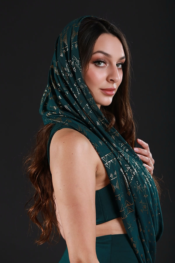 A woman with long, wavy brown hair models the Melodia Designs Alkimiya Infinity Scarf Malachite, a dark green hooded scarf with gold accents, paired with a matching outfit as she poses against a black background.