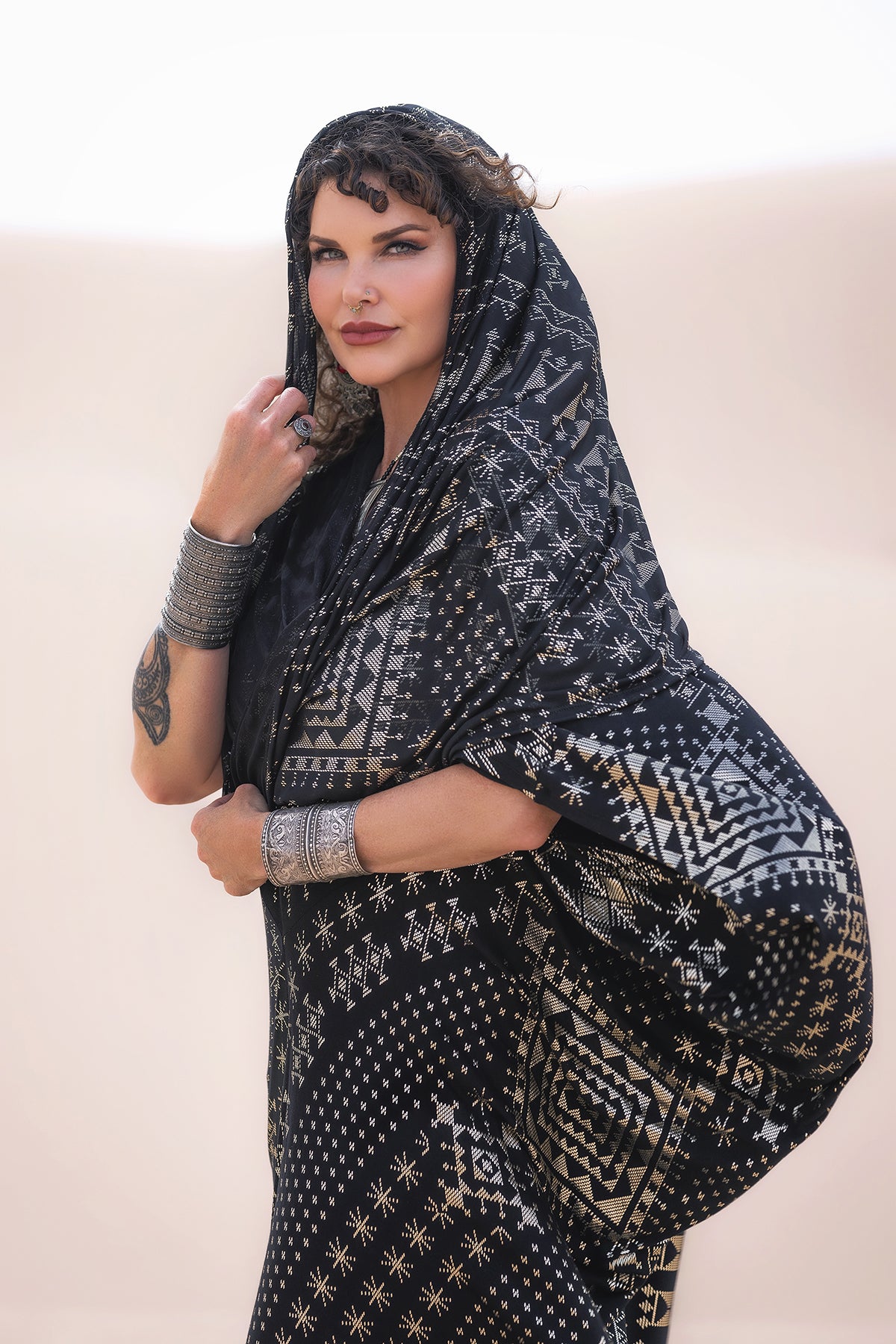 A woman in profile wears the Melodia Designs Alkimiya Infinity Scarf Obsidian—black and silver with a hood—styled as a statement piece. She pairs it with silver jewelry and stands before a soft, light backdrop.