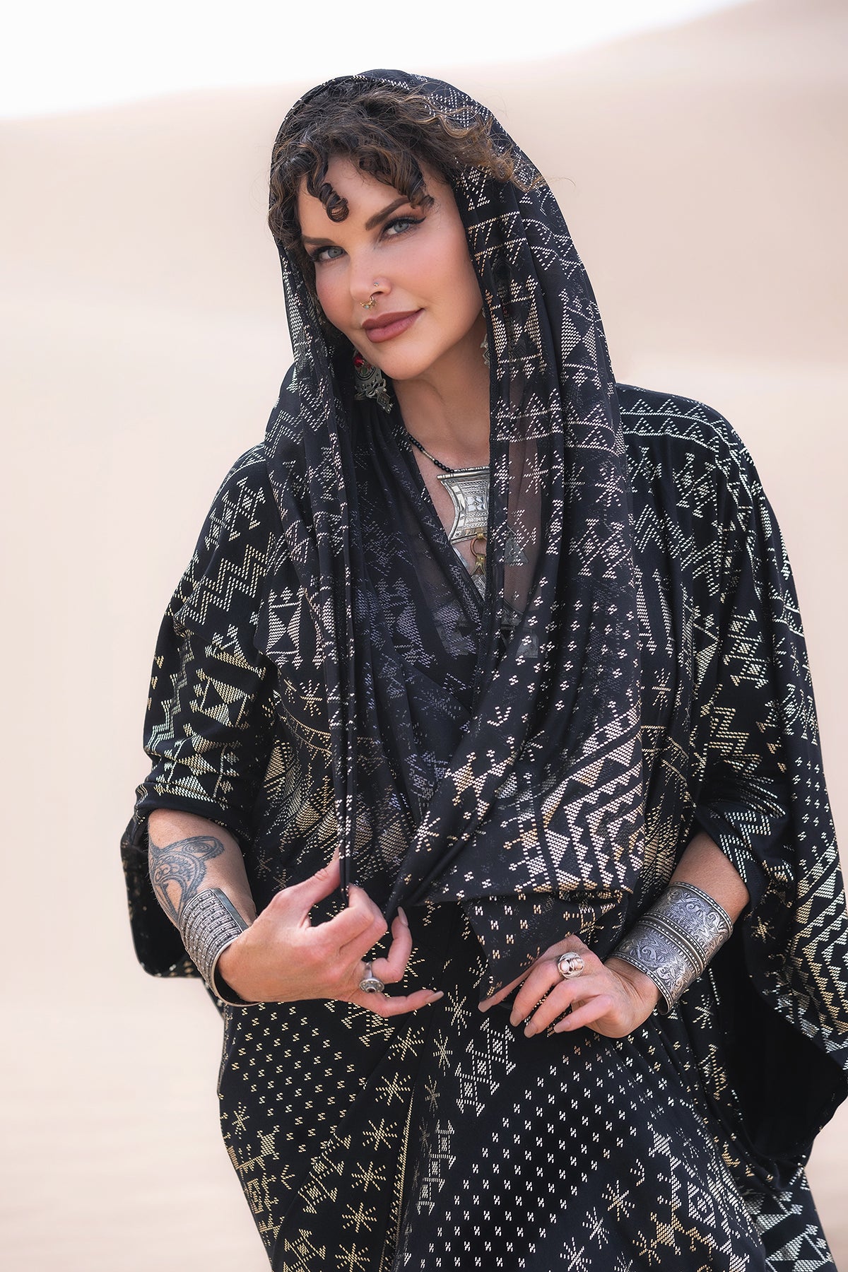 A woman in patterned black and gold attire stands in the desert, wearing Melodia Designs Alkimiya Infinity Scarf Obsidian. Adorned with silver jewelry and arm tattoos, she smiles softly at the camera.