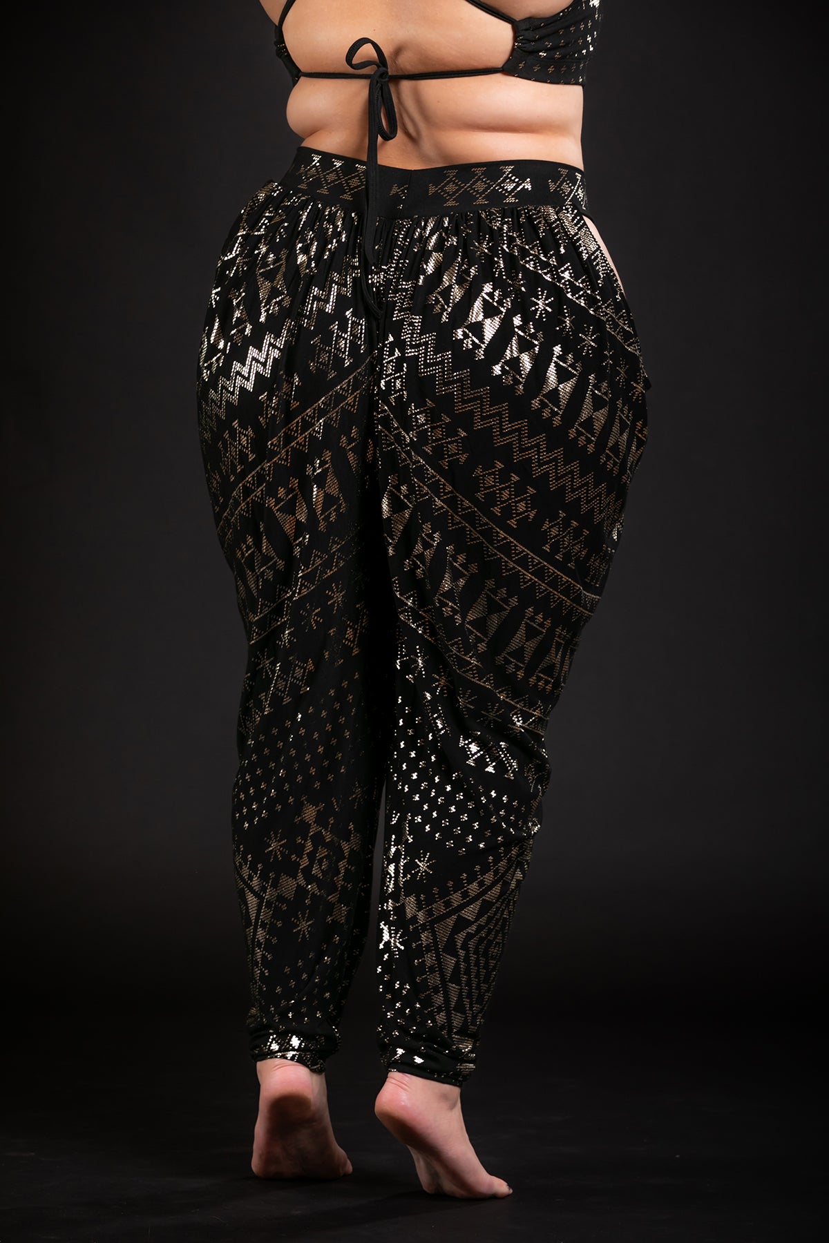 A person stands barefoot against a black background wearing Melodia Designs Alkimiya Isha Harem Pant Obsidian, featuring shimmering geometric and tribal motifs. The bamboo blend fabric is perfect for dance, with back and midriff visible under a matching crop top.